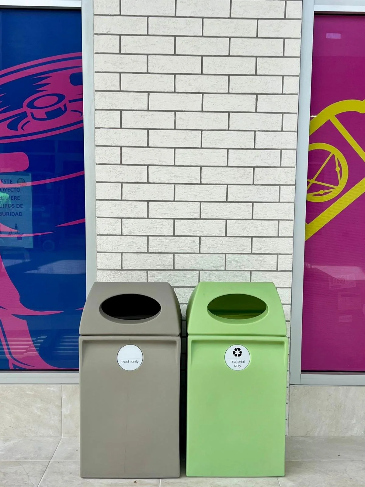Green and gray recycling bins placed side by side in front of a white brick wall, with colorful purple and blue signage on either side.