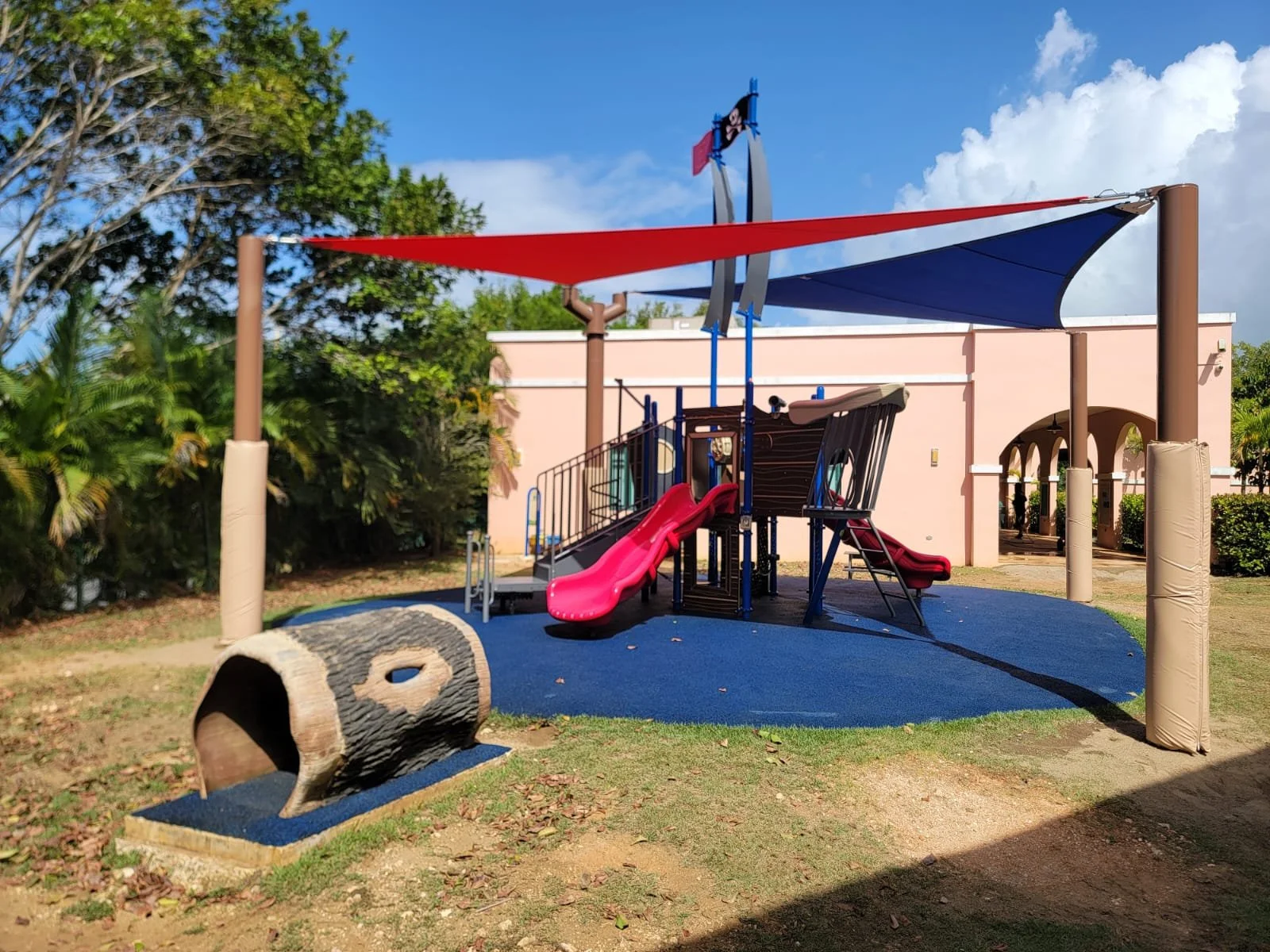 Colorful playground with red and blue shade sails, pink slides, and a wooden log sculpture on a blue rubberized surface, surrounded by greenery and a pink building in the background.