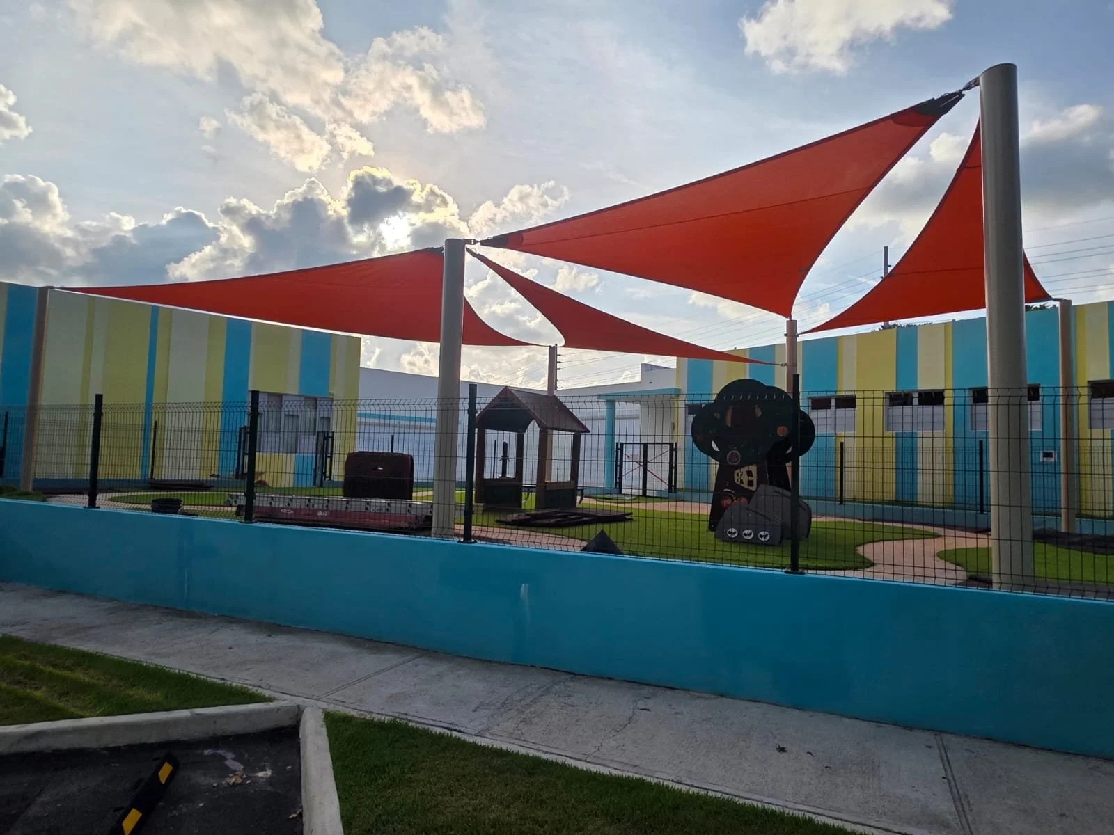 A playground area enclosed by a black fence features orange shade sails overhead, colorful striped walls, and playful structures including a tire and a train-like feature, with a sidewalk and a parking lot in the foreground under a partly cloudy sky.