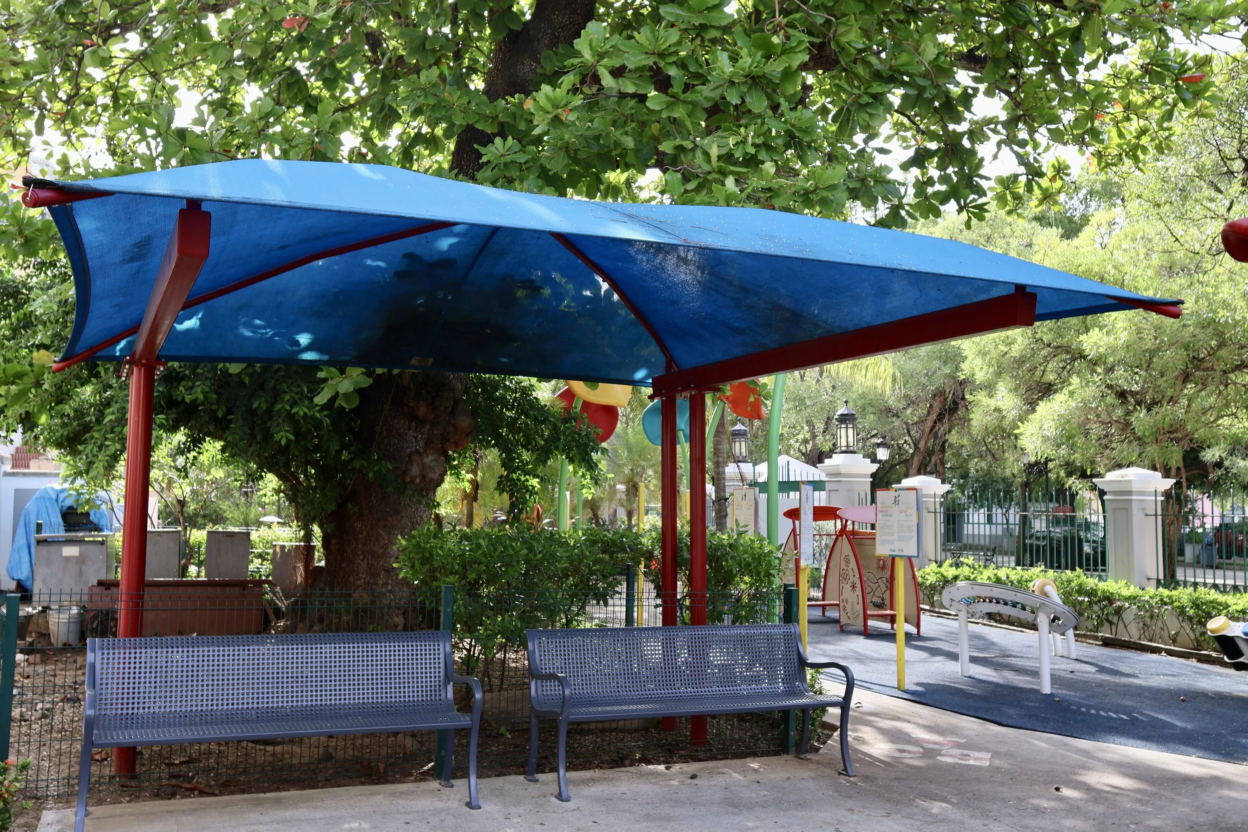 Public park area with blue benches, a shaded blue canopy, trees, and some colorful playground equipment in the background.