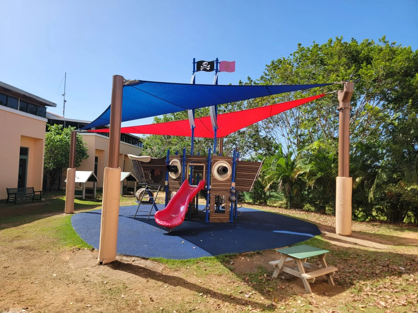Colorful playground with a red slide, pirate ship-themed play structure, blue and red shade sails, surrounded by green trees and a building in the background.