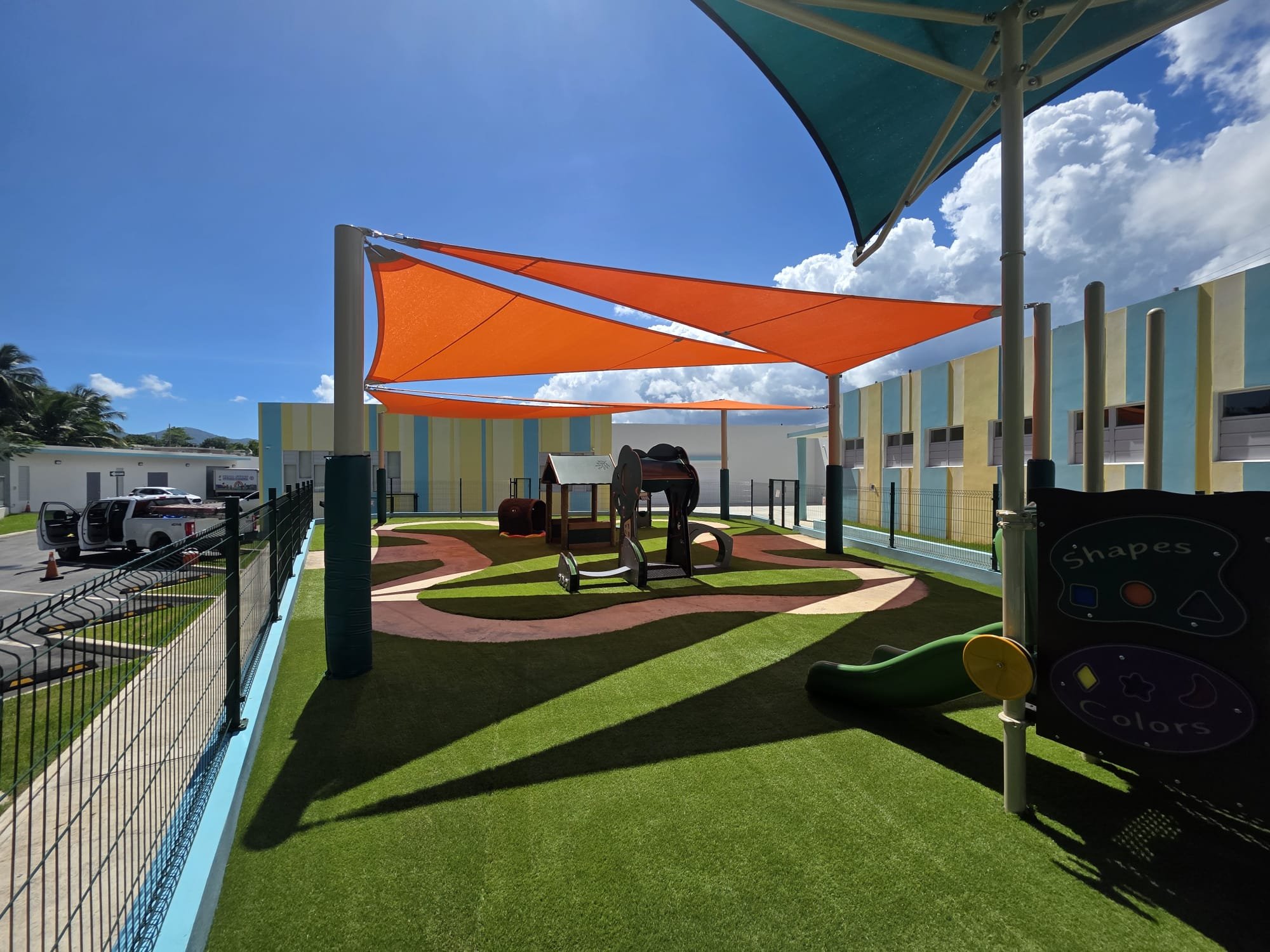 Colorful outdoor playground with shade sails, artificial grass, and play equipment, surrounded by a safety fence, on a sunny day.