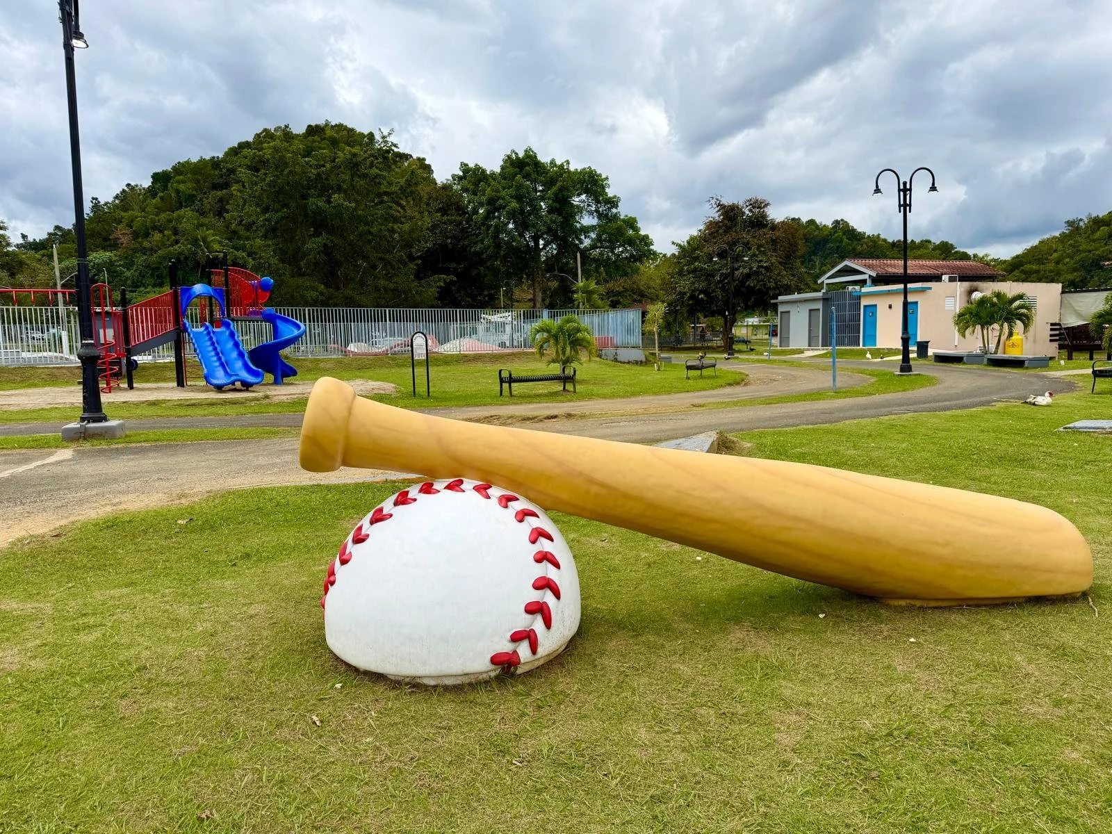 A baseball, a baseball bat, and a baseball glove arranged on grass in a park. In the background, there are playground equipment, benches, trees, a building, and cloudy sky.