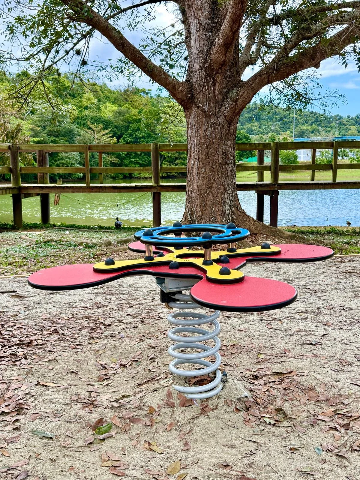 A colorful spring rider playground toy with red petals, yellow accents, a blue steering ring, and a metal spring base in a park near a pond, with a large tree and wooden fence in the background.