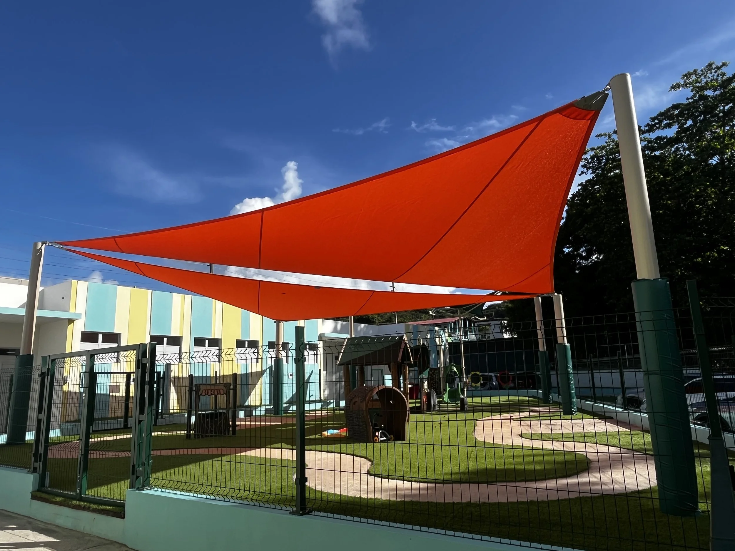 Colorful playground with a large, orange shade sail, enclosed by a fence, with a building in the background and a clear blue sky.