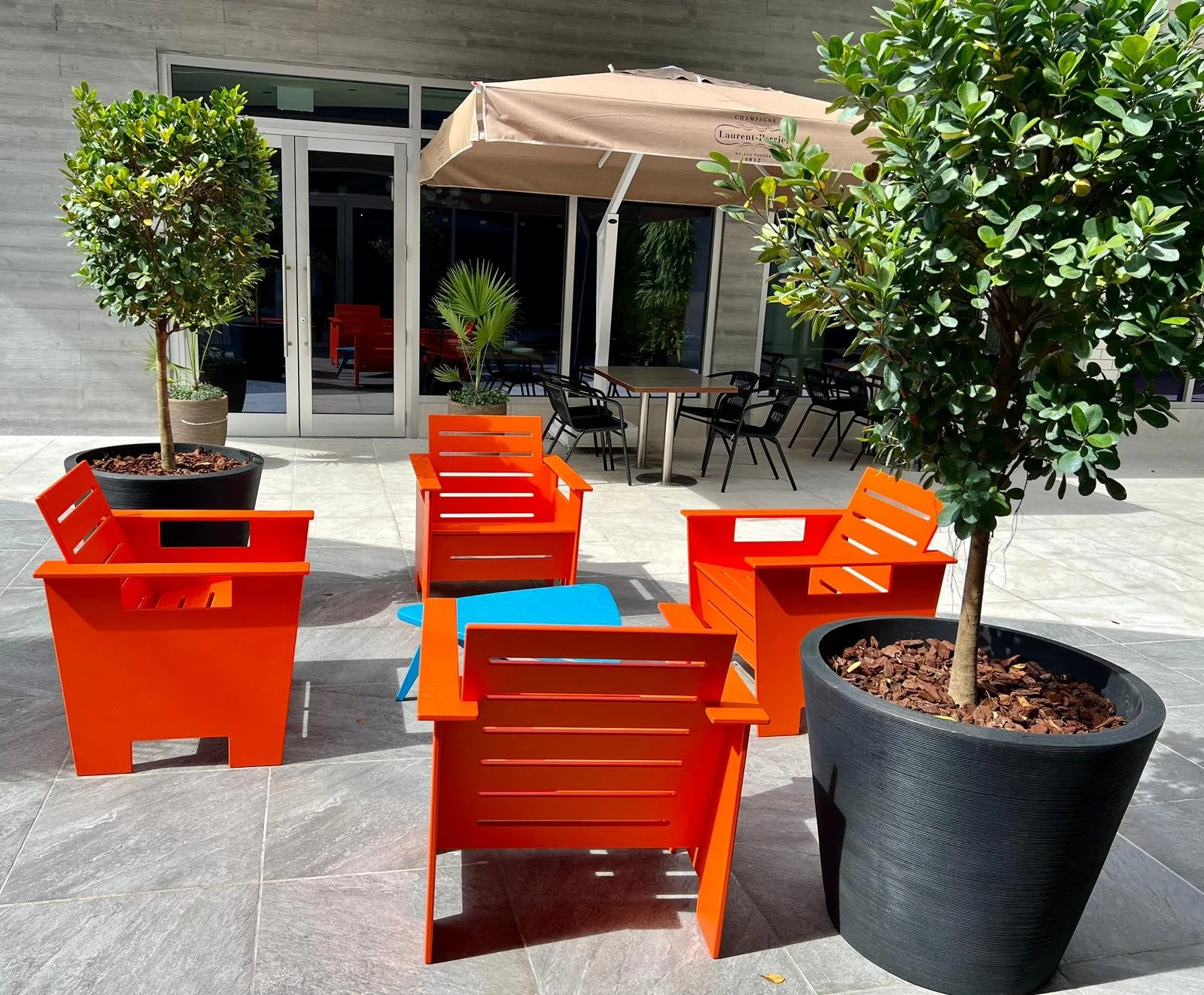An outdoor patio area with four bright orange chairs, two large potted trees, a small round blue table, and additional black chairs and tables under a beige umbrella.