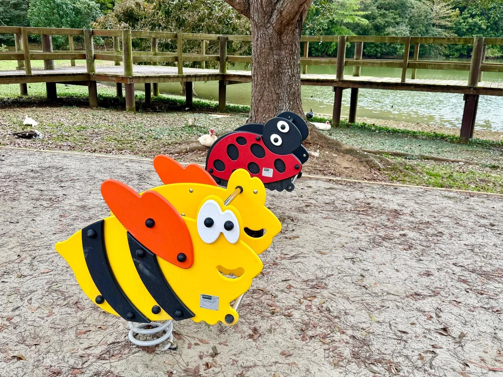 Colorful insect-shaped spring rider playground equipment in a park with a pond, wooden walkway, trees, ducks, and a tree in the background.