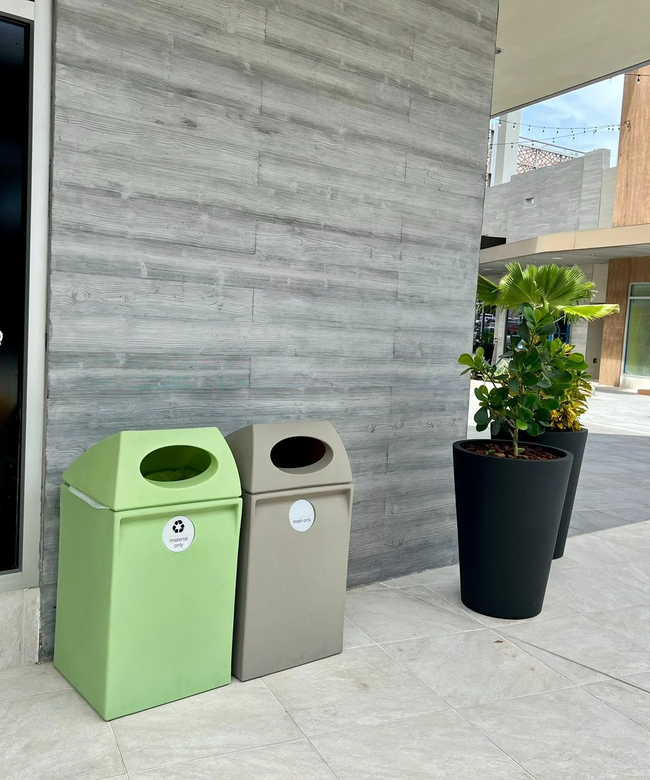 A lime green recycling bin and a gray trash can are placed against a gray wooden wall, with large potted plants nearby in an outdoor area.