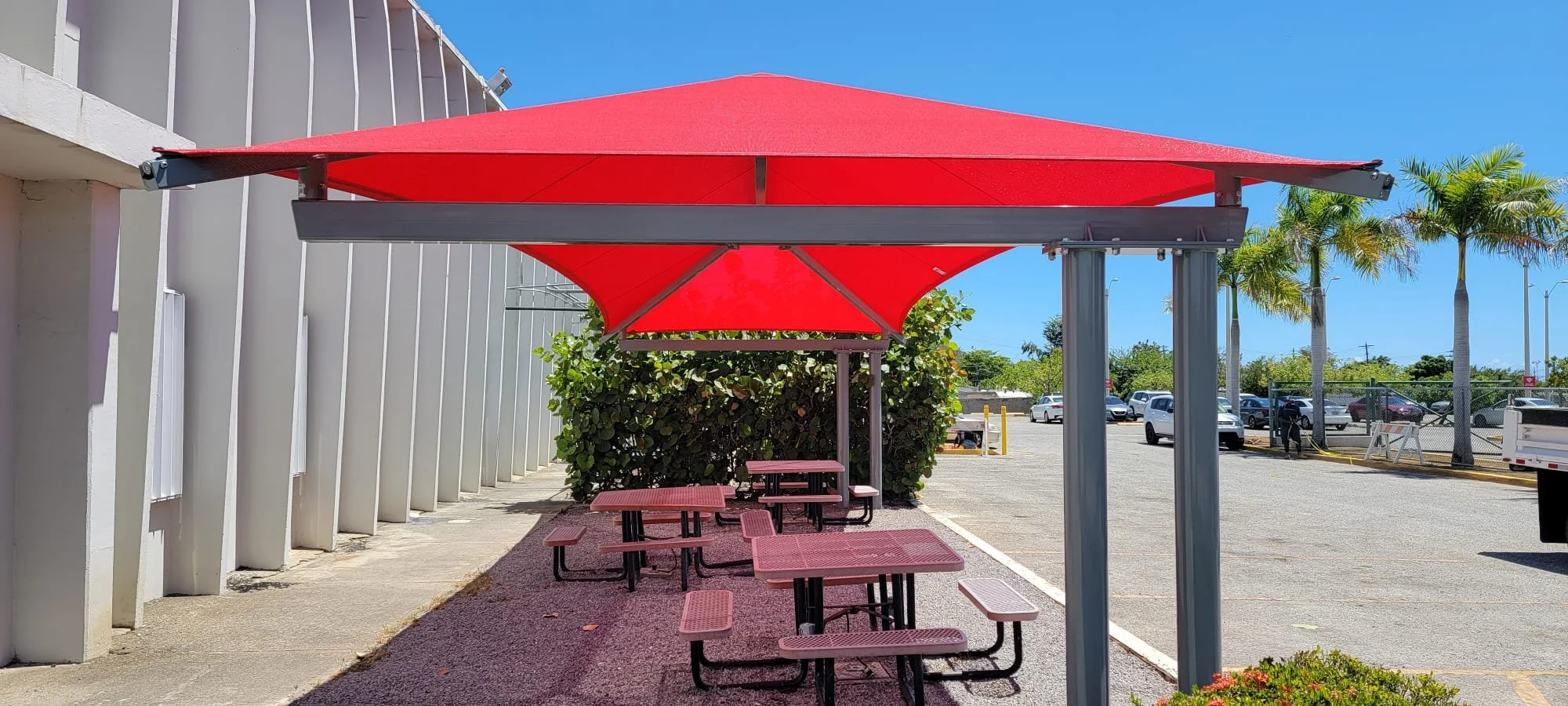 Outdoor seating area with red picnic tables and red shade canopy under a clear blue sky, near a parking lot with palm trees and a white building.