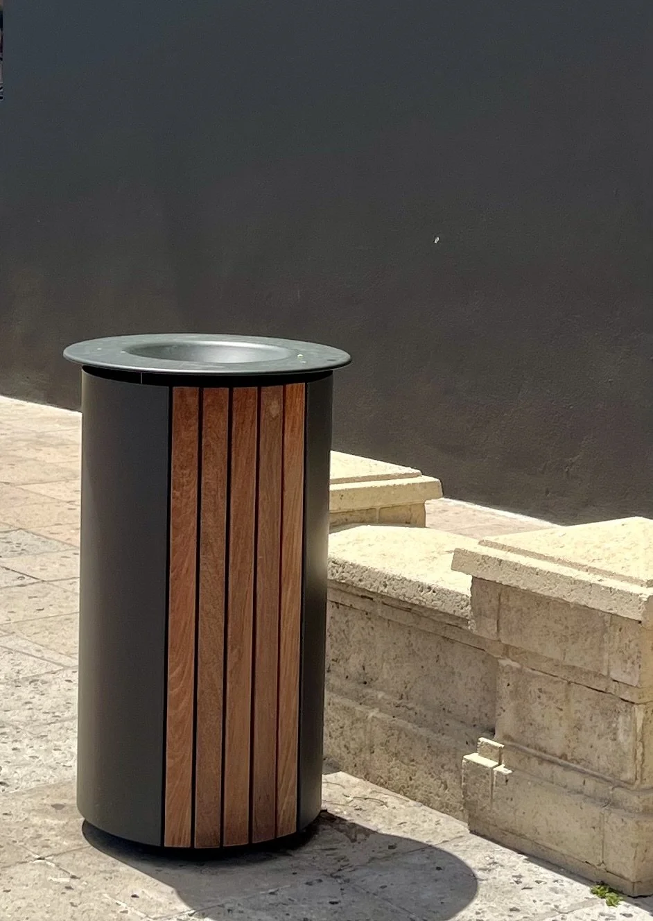 A cylindrical trash receptacle with a black metal top and bottom, and vertical wooden slats on the side, placed on a stone sidewalk near a black wall and a stone bench.