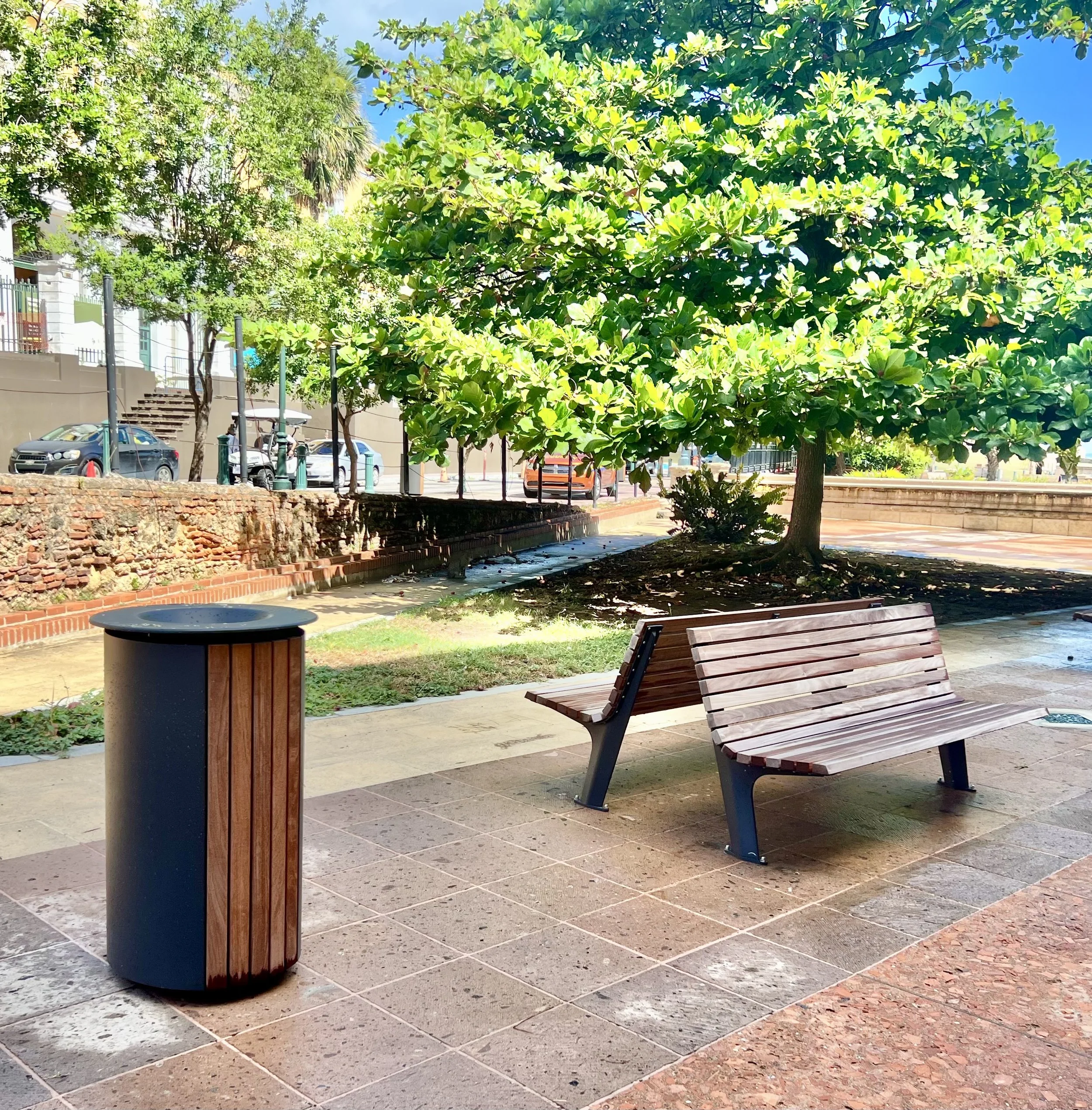 A park scene featuring a wooden bench, a trash can, and lush green trees with a sunny sky. In the background, there are parked cars, a building, and some stairs.