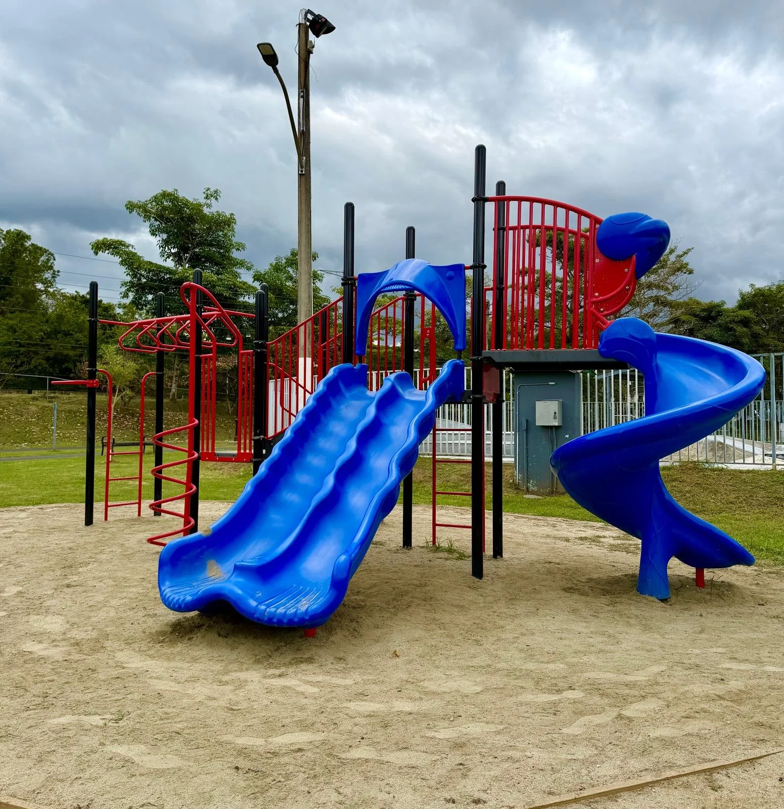 Children's playground with blue and red slides and climbing structures on sand surface, surrounded by green trees and cloudy sky.