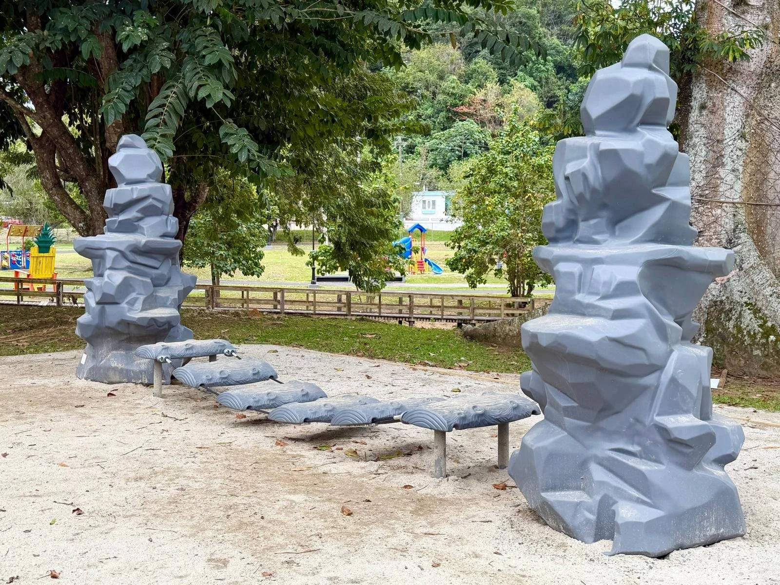 Modern sculpture playground structures in a park with trees and a playground slide in the background.