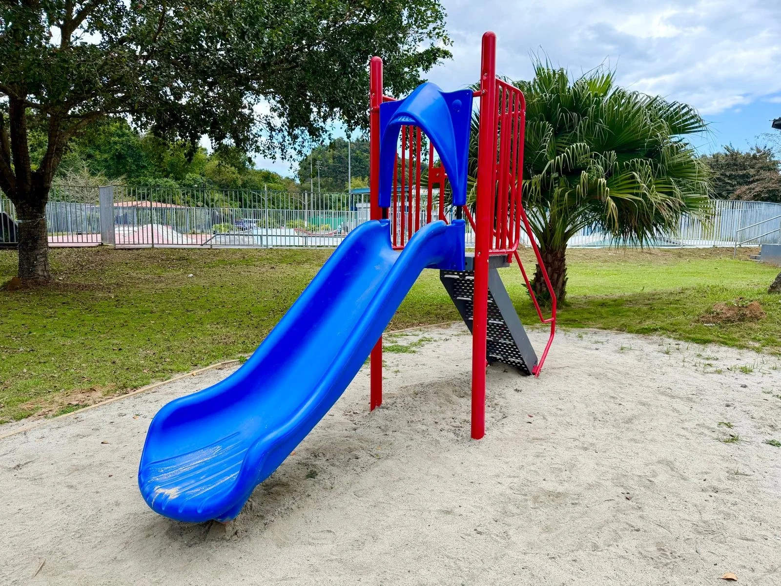 Colorful playground slide with a blue slide, red support bars, and a small staircase, situated on sandy ground within a park, with trees and a fence in the background.