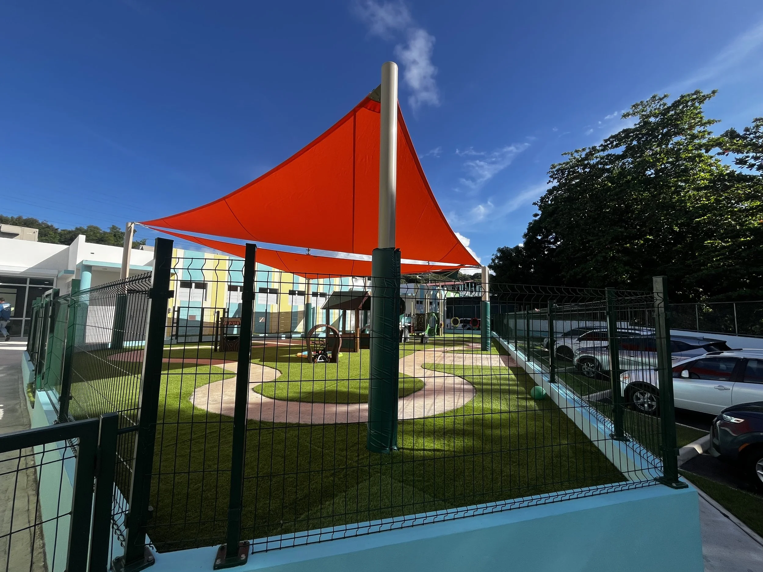 Colorful outdoor play area with a red shade sail, artificial grass, paved pathways, and parking lot with cars, surrounded by a black metal fence and trees, under a clear blue sky.