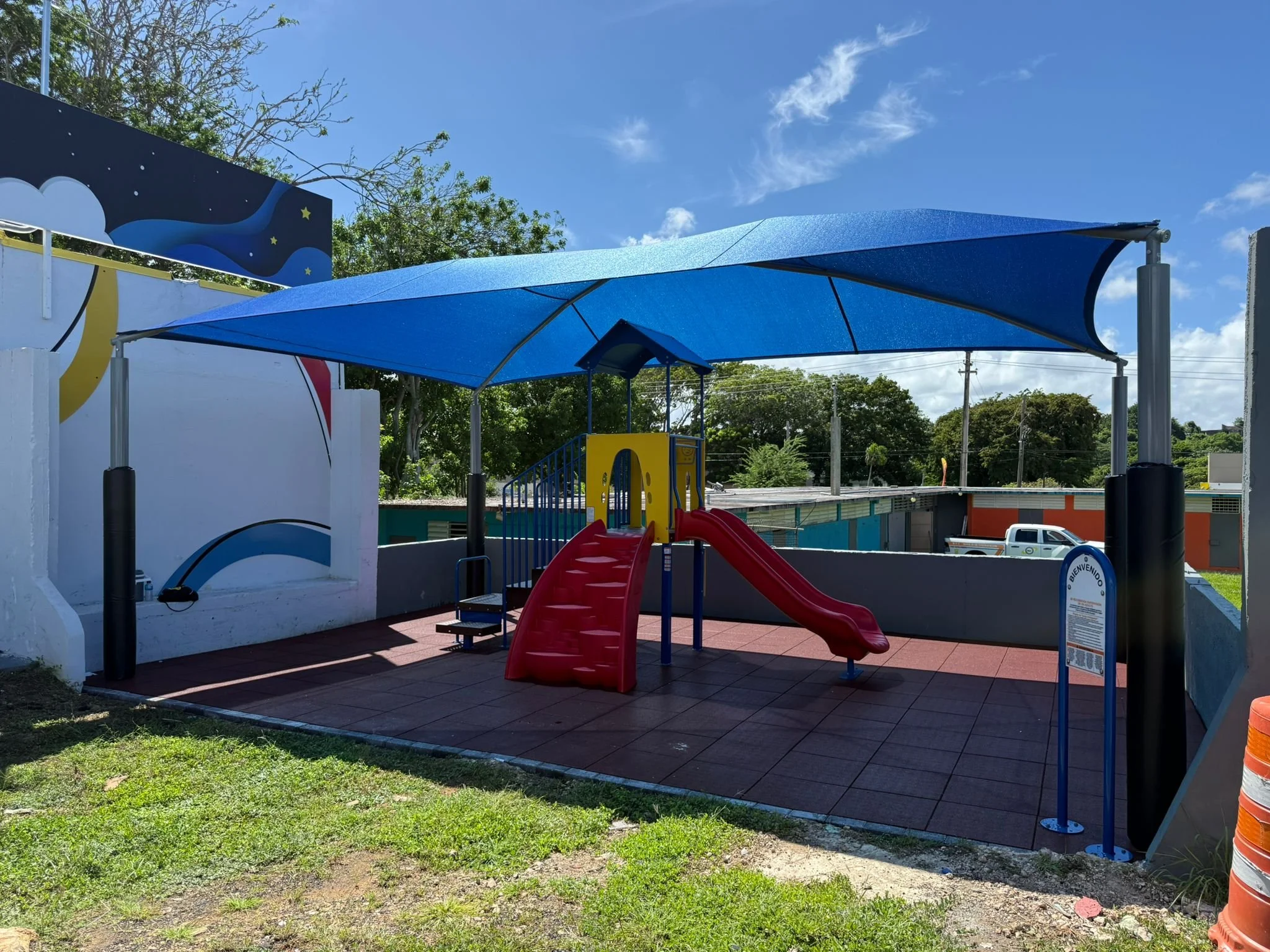 A playground with a red slide, a blue shade canopy, and a playground structure with stairs, set in an area with tiles and grass, under a partly cloudy sky.