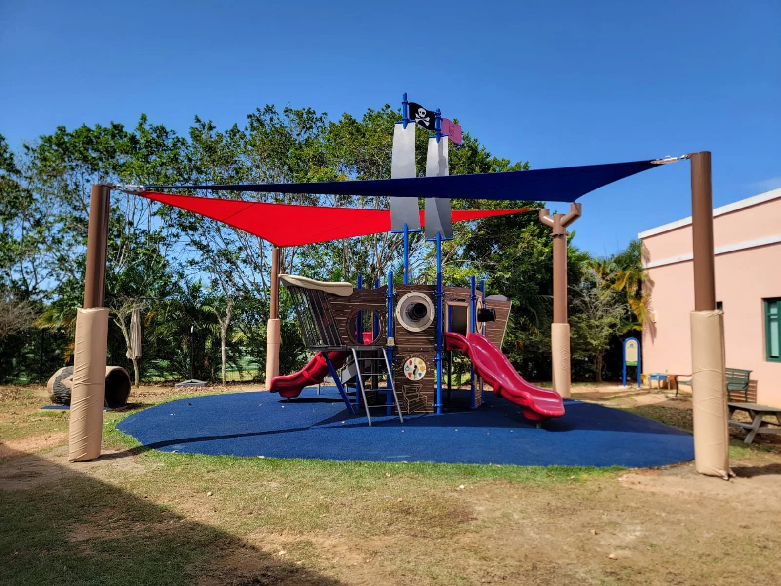 Colorful playground with pirate ship structure, slides, and shade sails, in a park with trees and a building nearby.
