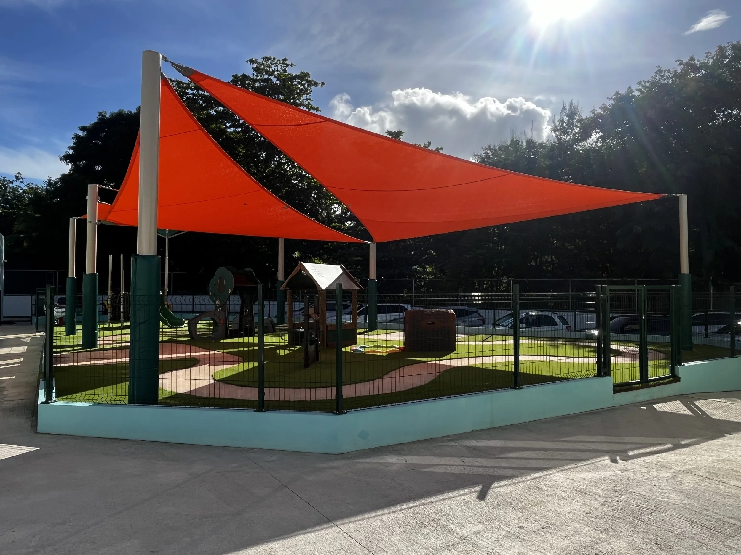 Colorful outdoor playground with orange shade sails, small tunnels, and grassy play area, fenced in with parking lot nearby.