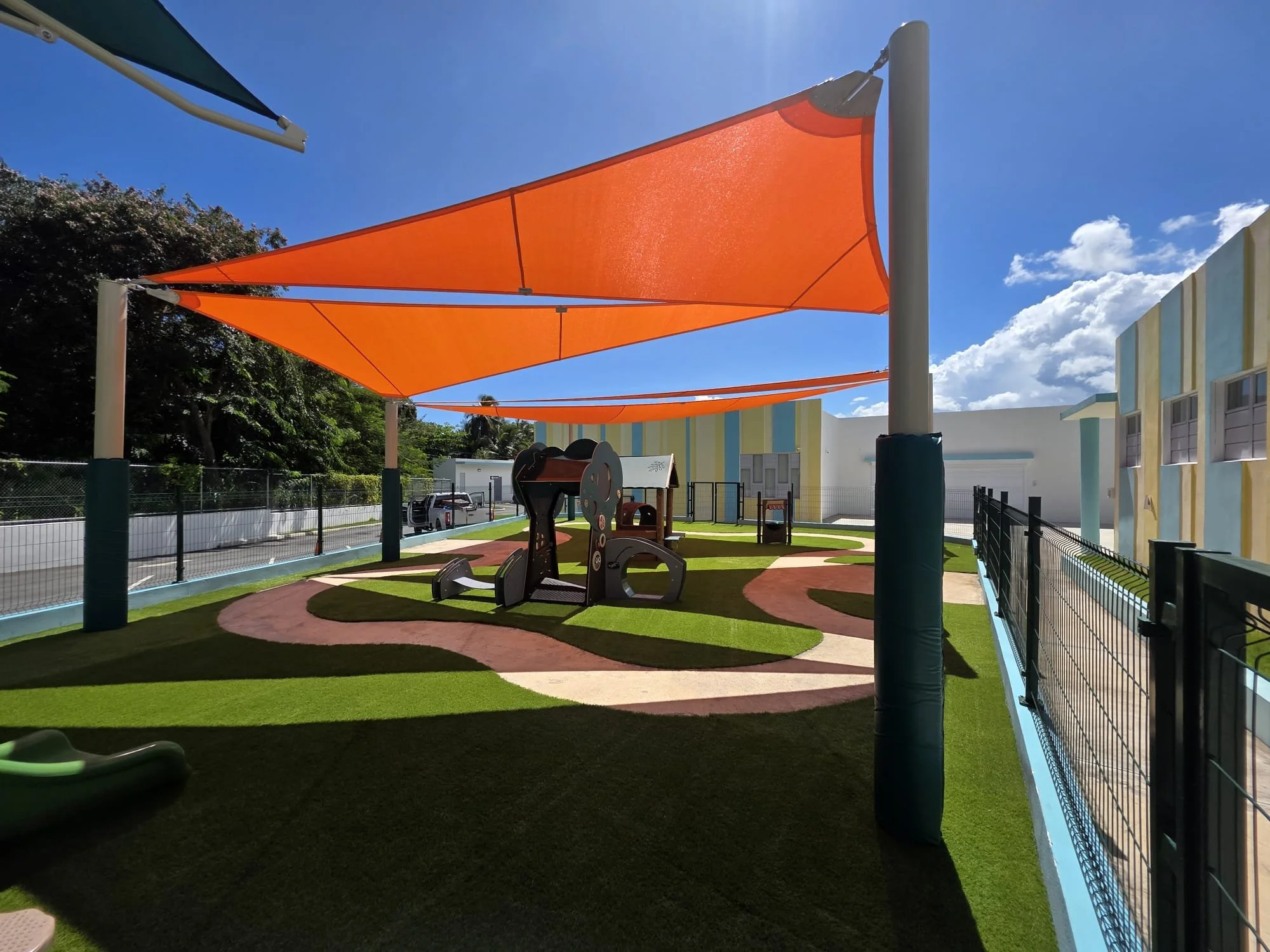 Colorful outdoor playground with orange shade sails, play equipment, artificial grass, white building with colorful stripes, and clear blue sky.