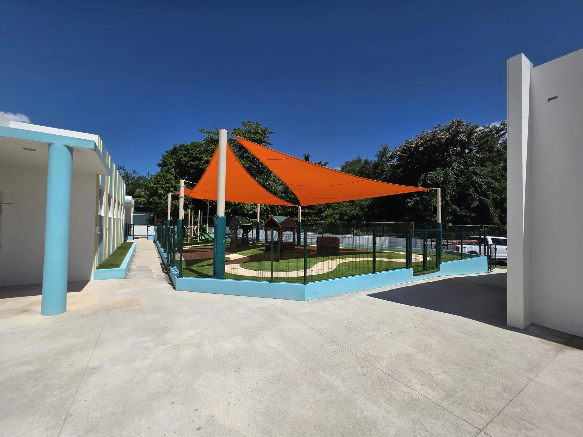 Childrens playground with orange shade sails, surrounded by a black fence, in a school or daycare facility, with a white building to the left and trees in the background under a clear blue sky.