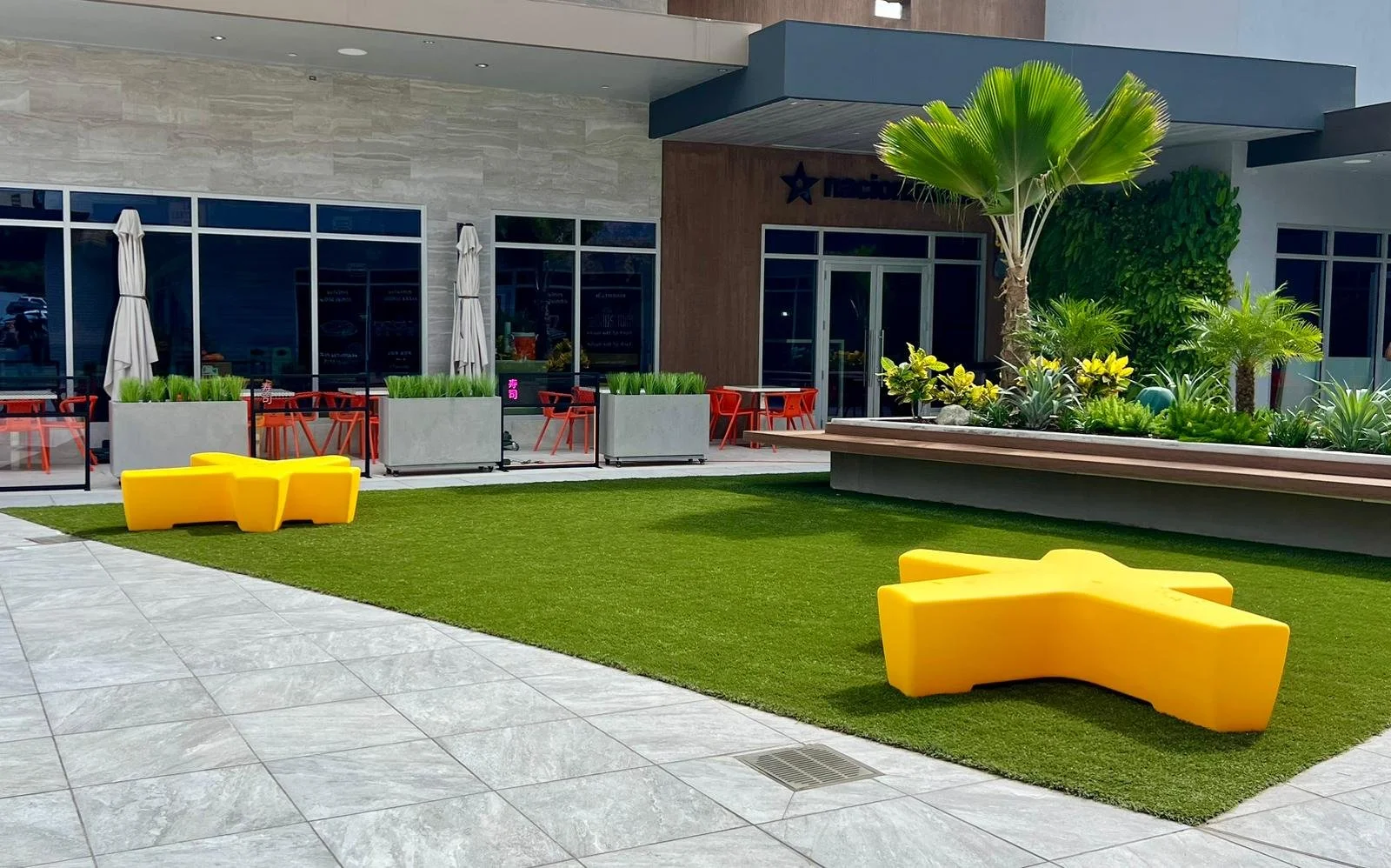 Outdoor patio with modern yellow chairs on artificial grass and a landscaped garden with palm trees in front of a building with large glass windows and patio seating.