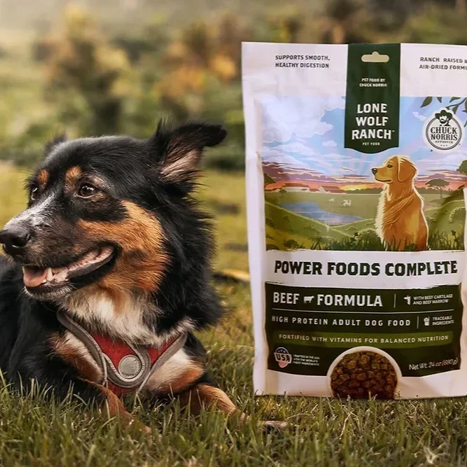A dog sitting on grass next to a bag of Lone Wolf Ranch Power Foods Complete beef formula dog food outdoors with a scenic landscape background