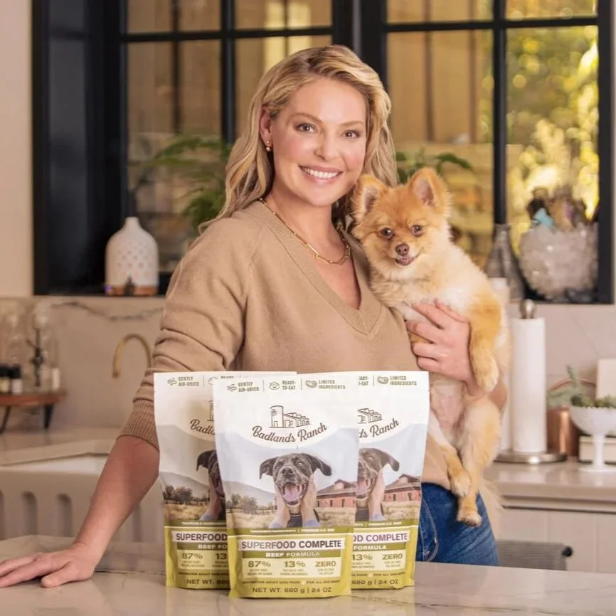 A smiling woman in a beige sweater holding a small fluffy dog in a kitchen. On the counter in front of her are three bags of Badlands Ranch dog food.