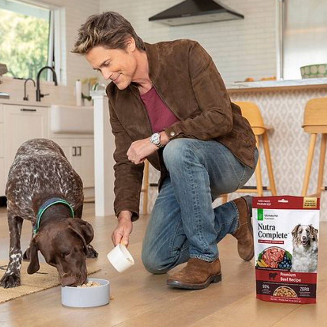 A man in a brown jacket and jeans kneels on the floor, feeding dog food to a brown dog with a green collar using a scoop. There is a bag of Nutra Complete dog food nearby in the kitchen.