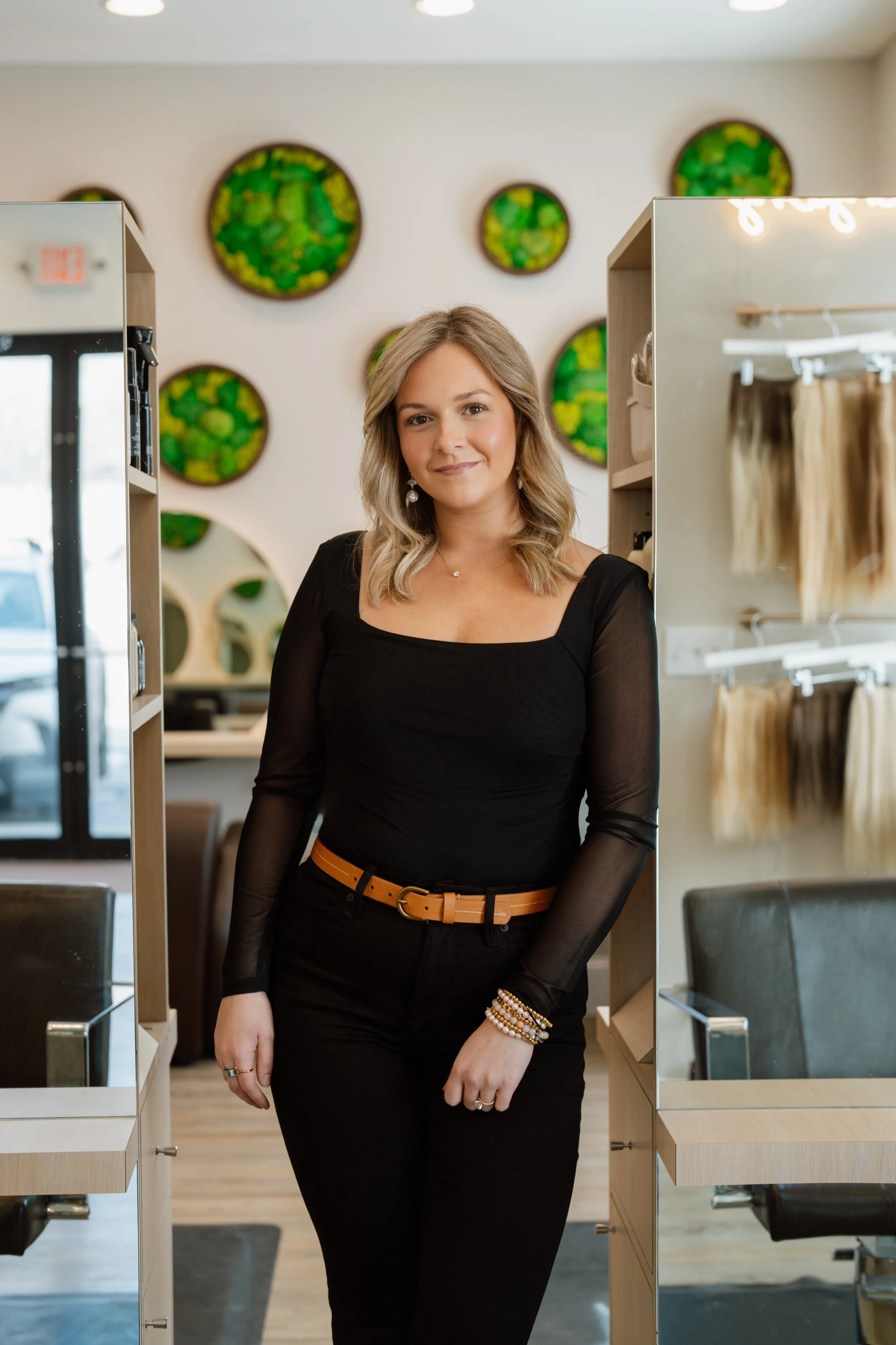 A woman with blonde hair wearing a black dress and a tan belt standing in a store with display shelves and green circular wall art.