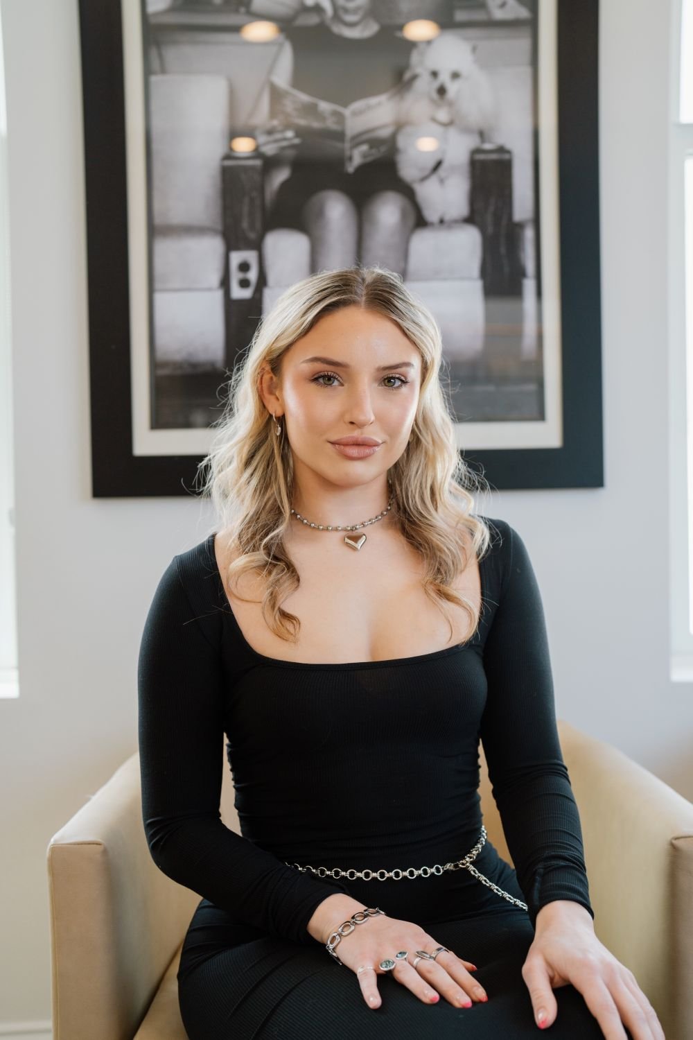 A young woman with blonde, wavy hair, wearing a black long-sleeve top, silver jewelry including a necklace, rings, bracelets, and an accessory at the waist, sitting on a beige couch in a well-lit room with white walls. Behind her, there is a black and white framed photograph of a dog sitting on a sofa with a person.