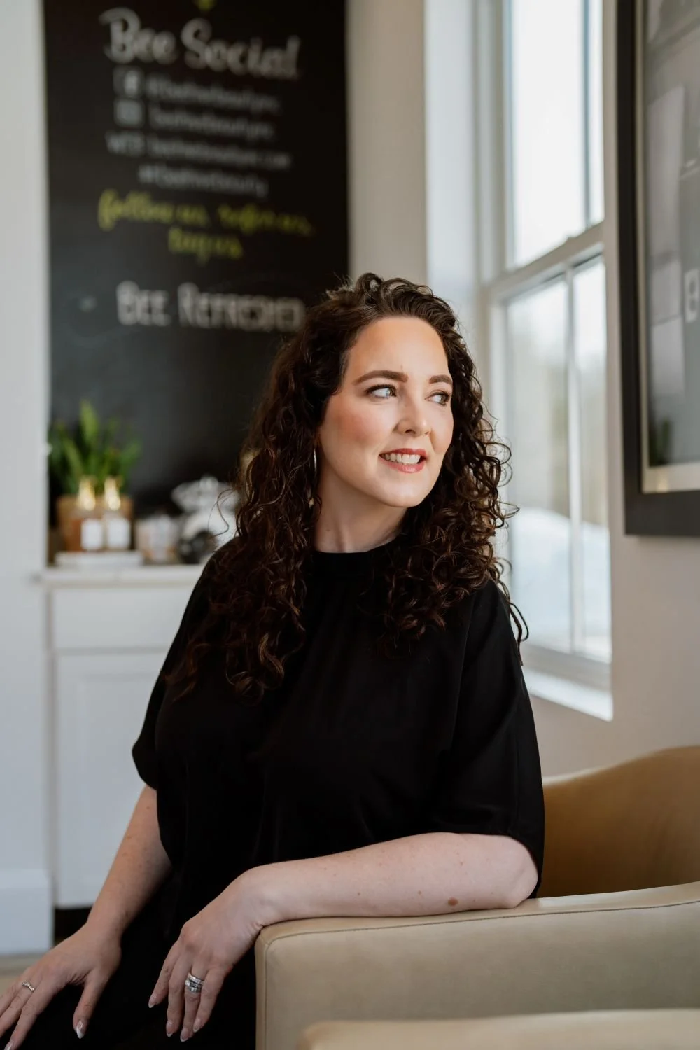 A woman with curly brown hair, wearing a black top, sitting on a beige chair in a cafe, looking to her left. In the background, there is a blackboard with partially visible text and some potted plants.