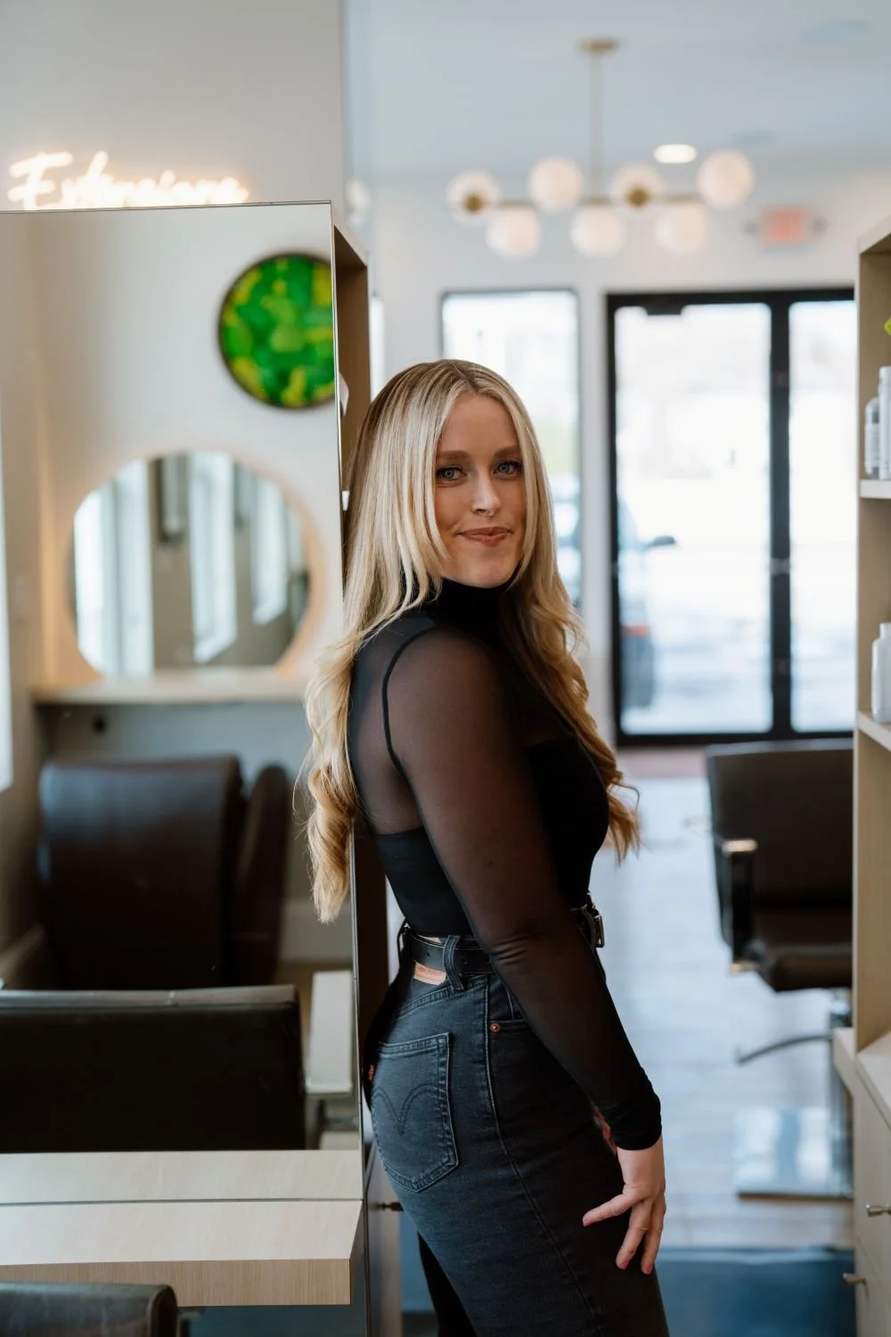 A young woman with long blonde hair wearing a black sheer long sleeve top and black jeans standing inside a modern salon or hair studio.