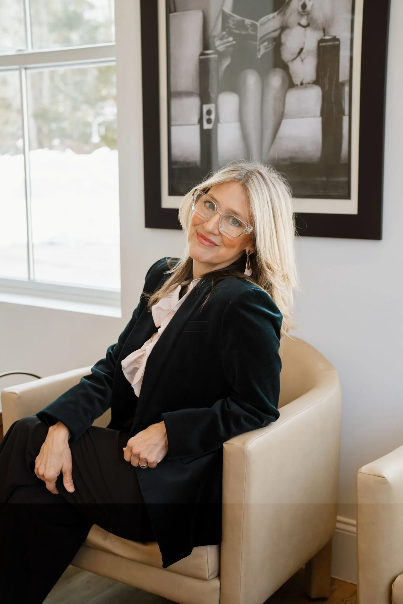 A middle-aged woman with blonde hair, wearing glasses, a black blazer, and a pink scarf, sitting in a cream-colored armchair in a well-lit room with large windows. Behind her is a black and white framed photograph of a woman's legs and torso.