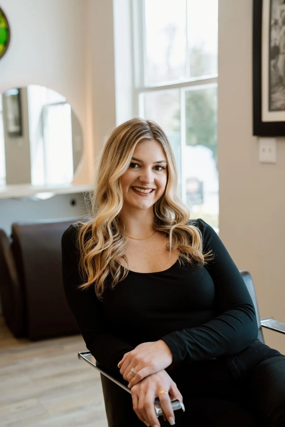 A woman with blonde, wavy hair smiling while sitting in a salon chair near a window, wearing a black long-sleeve top and jewelry.
