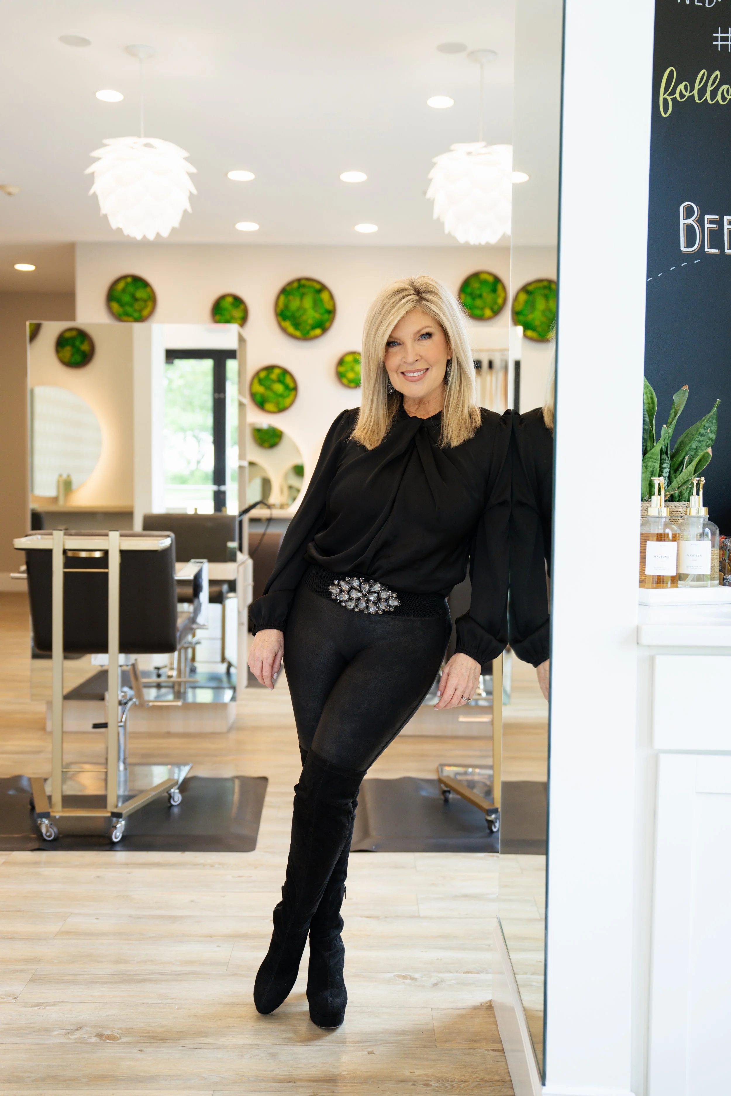 A woman with blonde hair wearing a black dress with a decorative belt, standing and smiling inside a modern salon with white walls, circular green wall decor, and bright lighting.