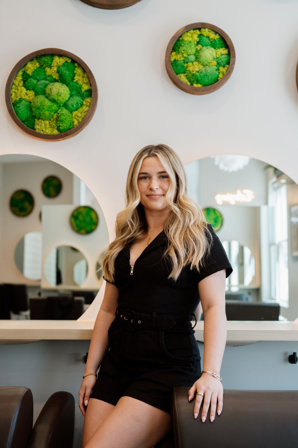 A young woman with long blonde hair standing in a modern interior with circular moss wall art.