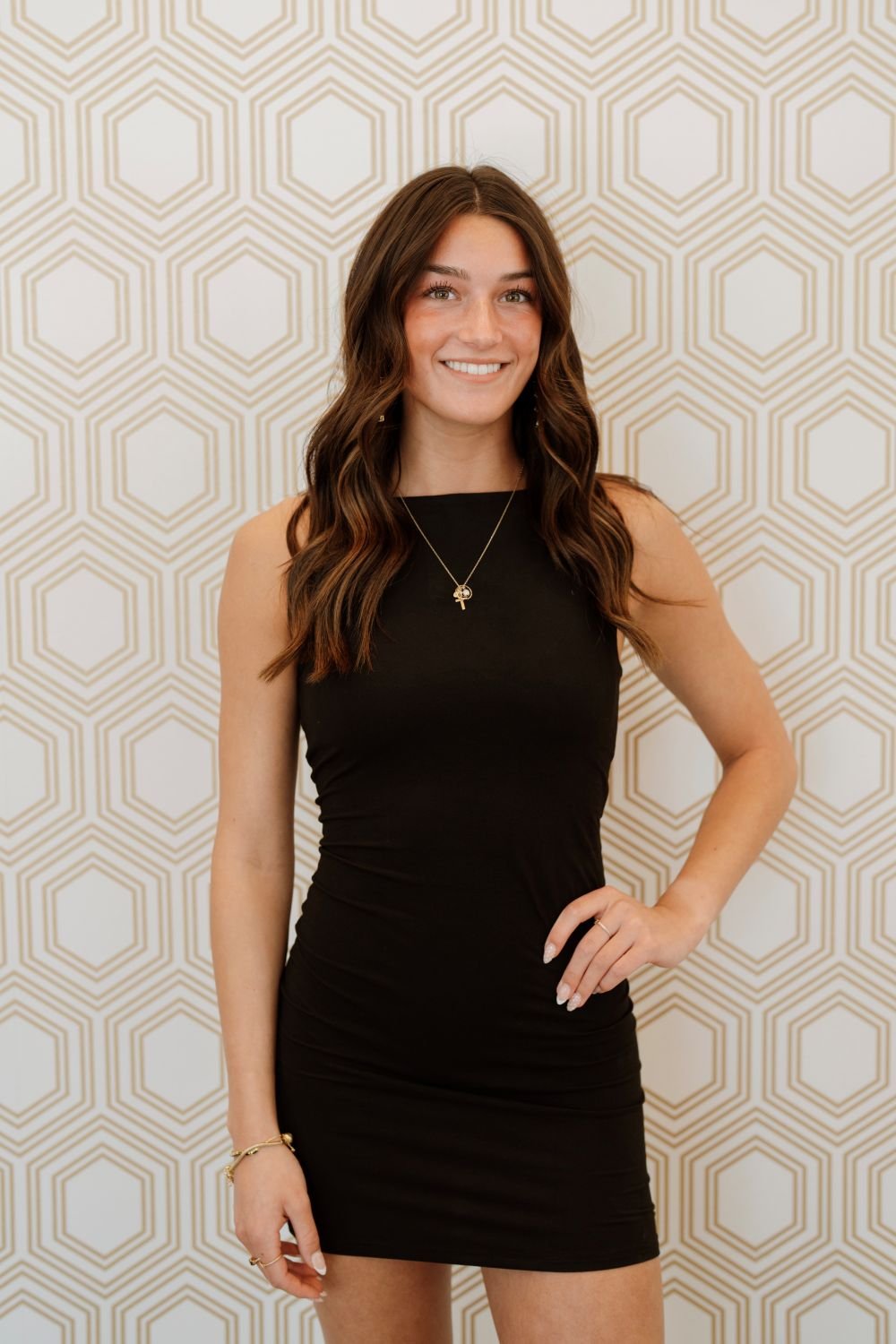A young woman in a sleeveless black dress standing against a geometric patterned wall, smiling with her hand on her hip, wearing a necklace and bracelets.