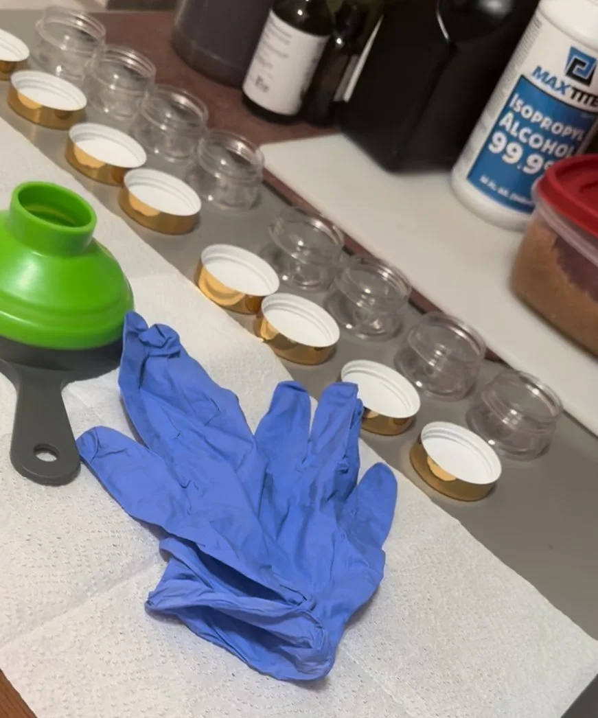 A table with small glass jars, some with white and gold lids, a pair of blue gloves, a green pill crusher, and various bottles including isopropyl alcohol, indicating preparation for medication or sample handling.