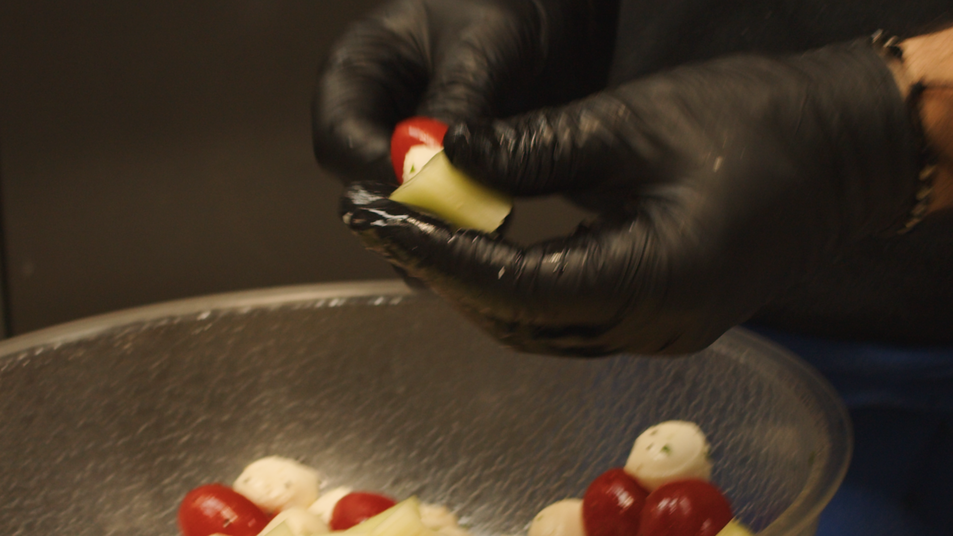 A person wearing black gloves preparing a salad with cherry tomatoes, mozzarella balls, and green onions in a glass bowl.