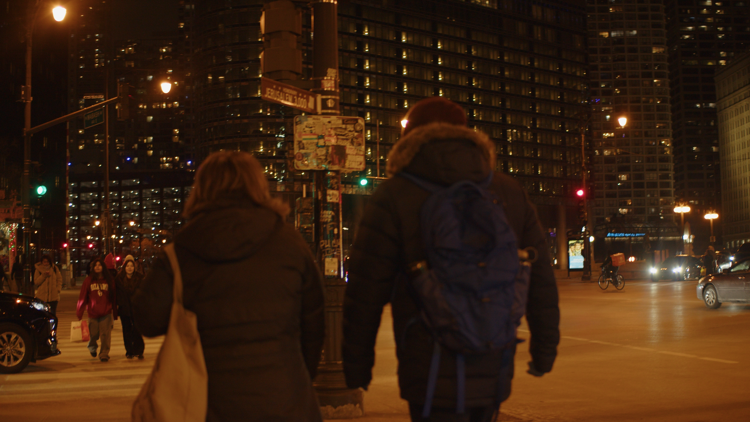 Night scene of a city street intersection with pedestrians crossing, cars, and tall buildings illuminated in the background.