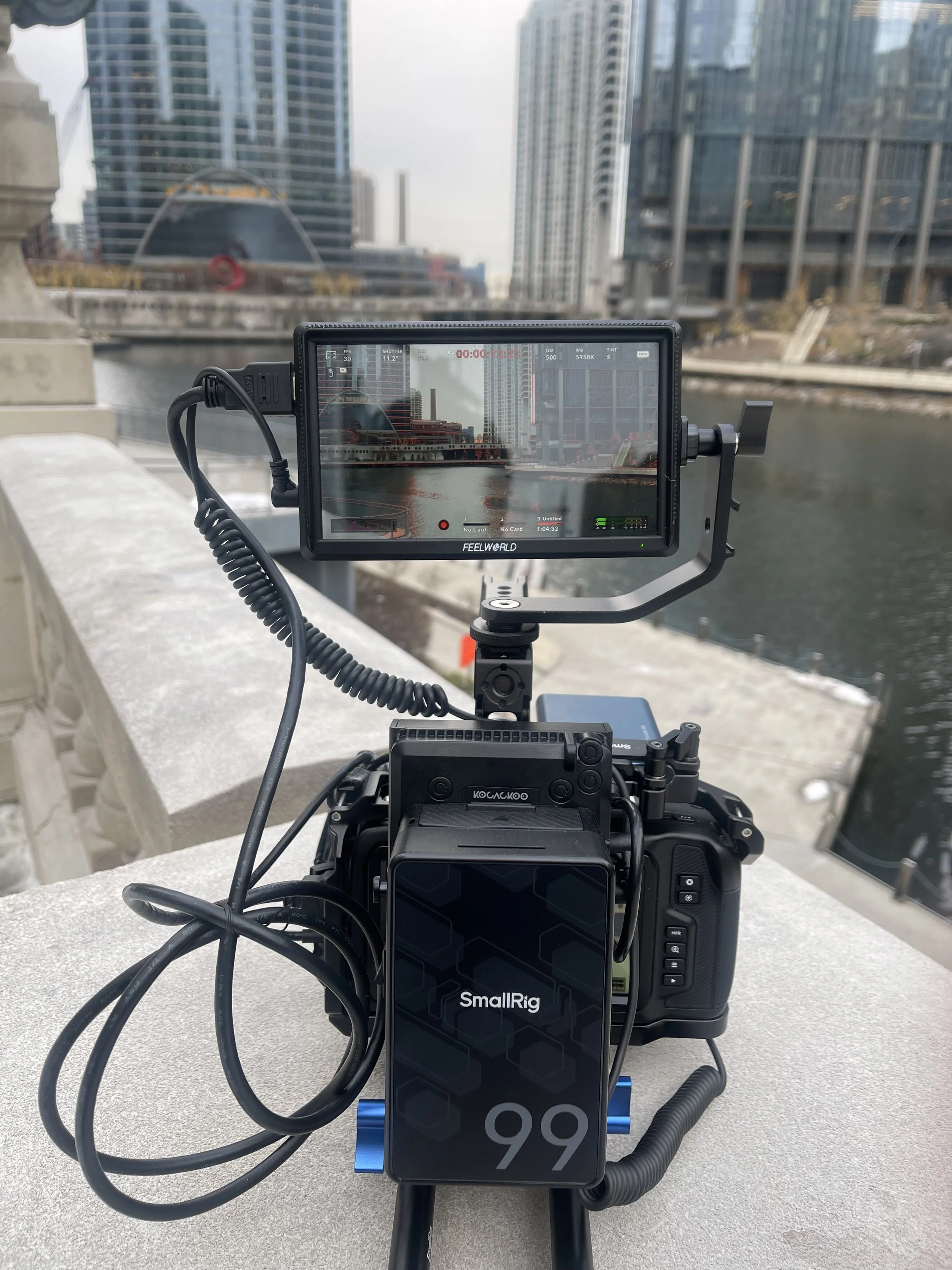 A professional camera setup with an external monitor on a tripod, positioned on a bridge over a river, with city buildings in the background.