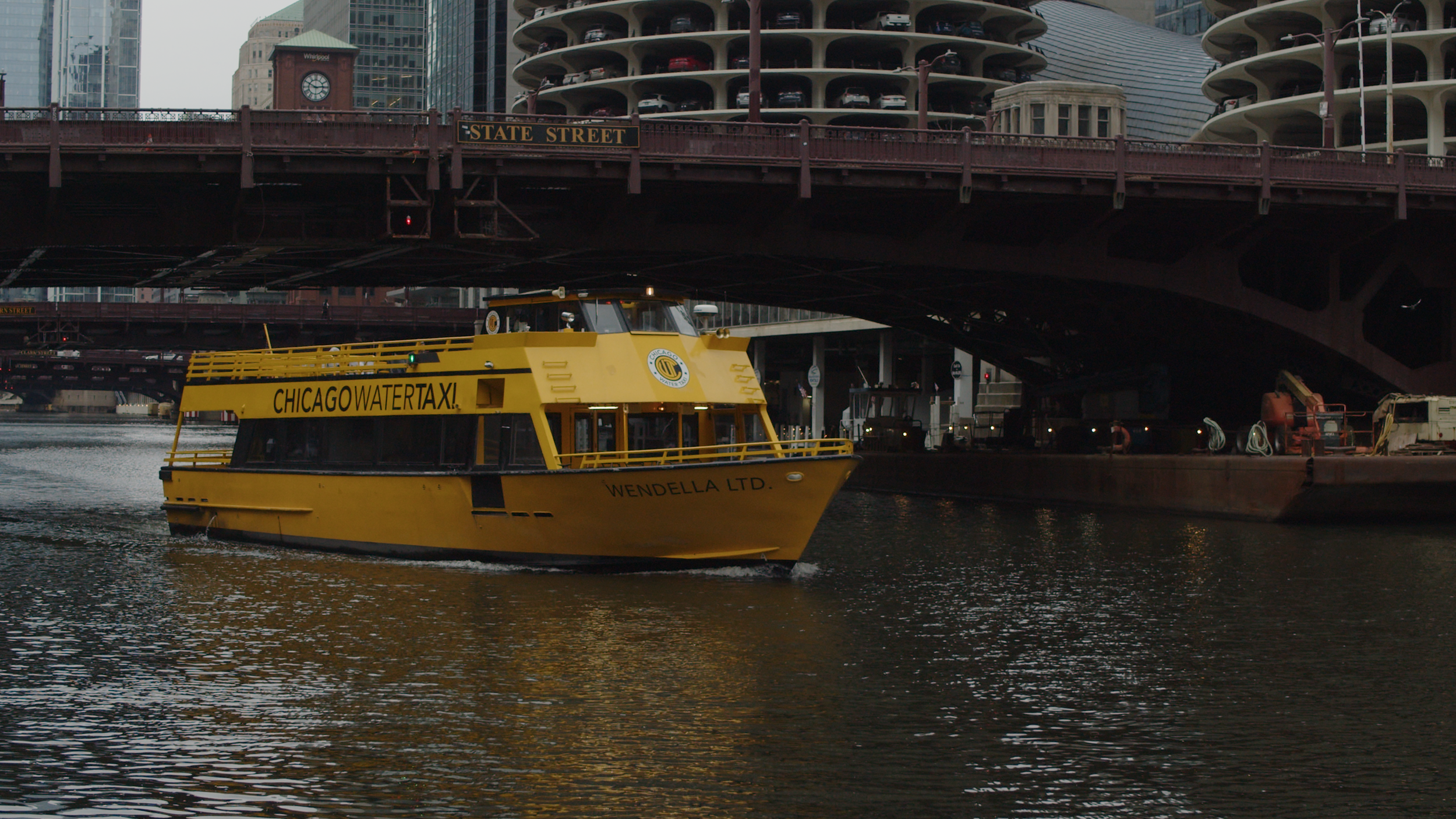 A yellow Chicago water taxi boat named 'Wendella Ltd.' sailing under a bridge on a river with city buildings in the background.