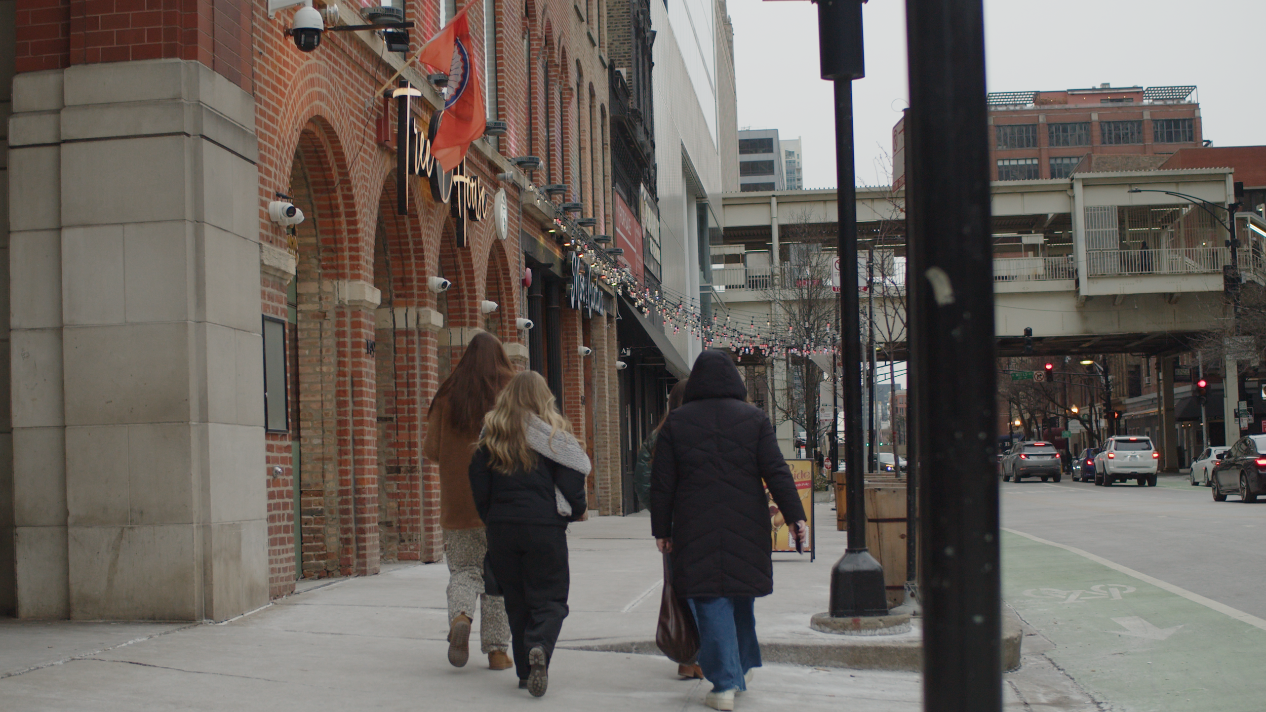 People walking on city sidewalk near brick building with decorations, cars on street, and overpass in the background.