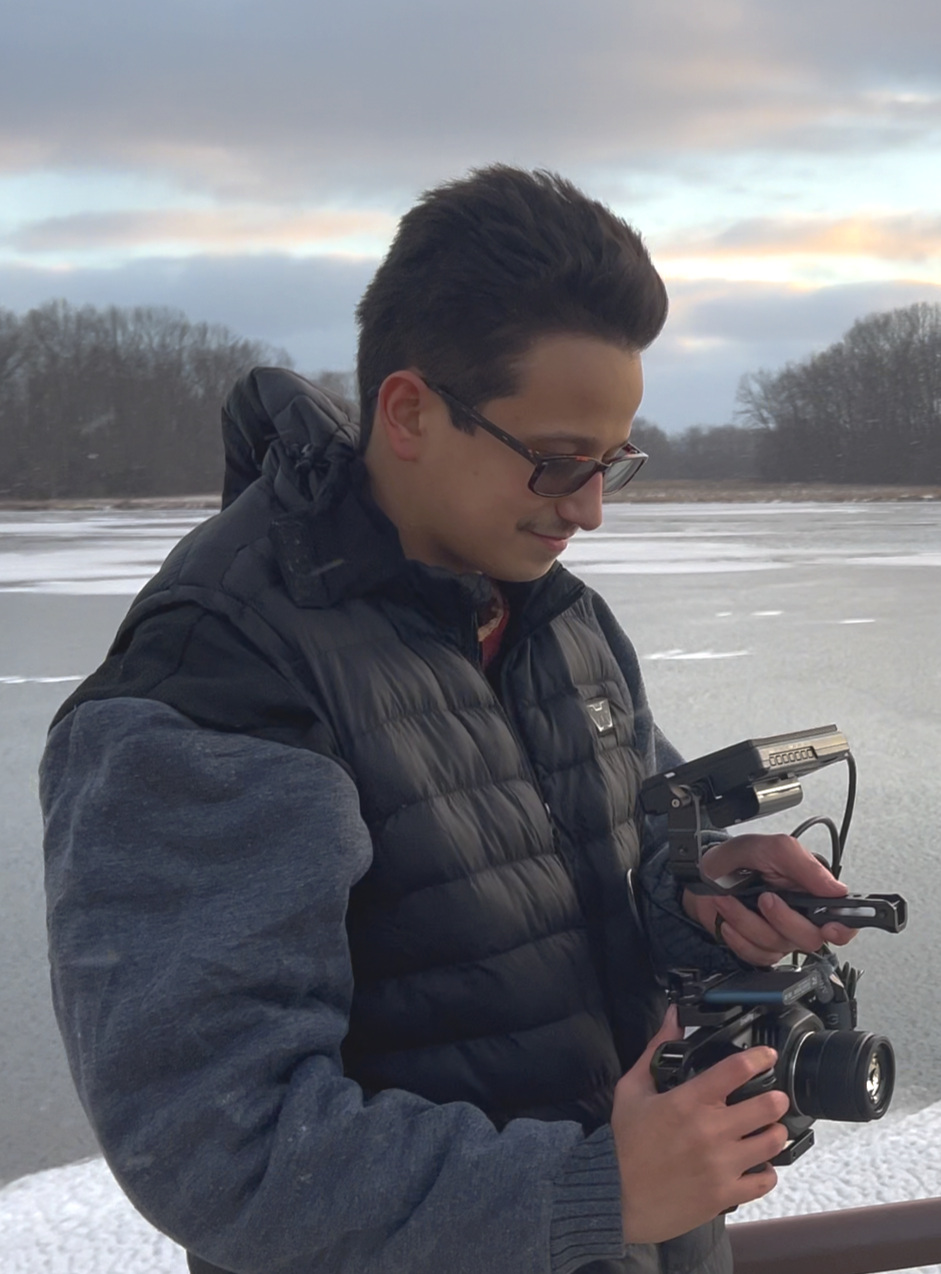 A young man wearing glasses, a black puffy vest, and a gray sweatshirt using a camera with a mounted camera stabilizer outdoors near a frozen body of water with trees in the background.