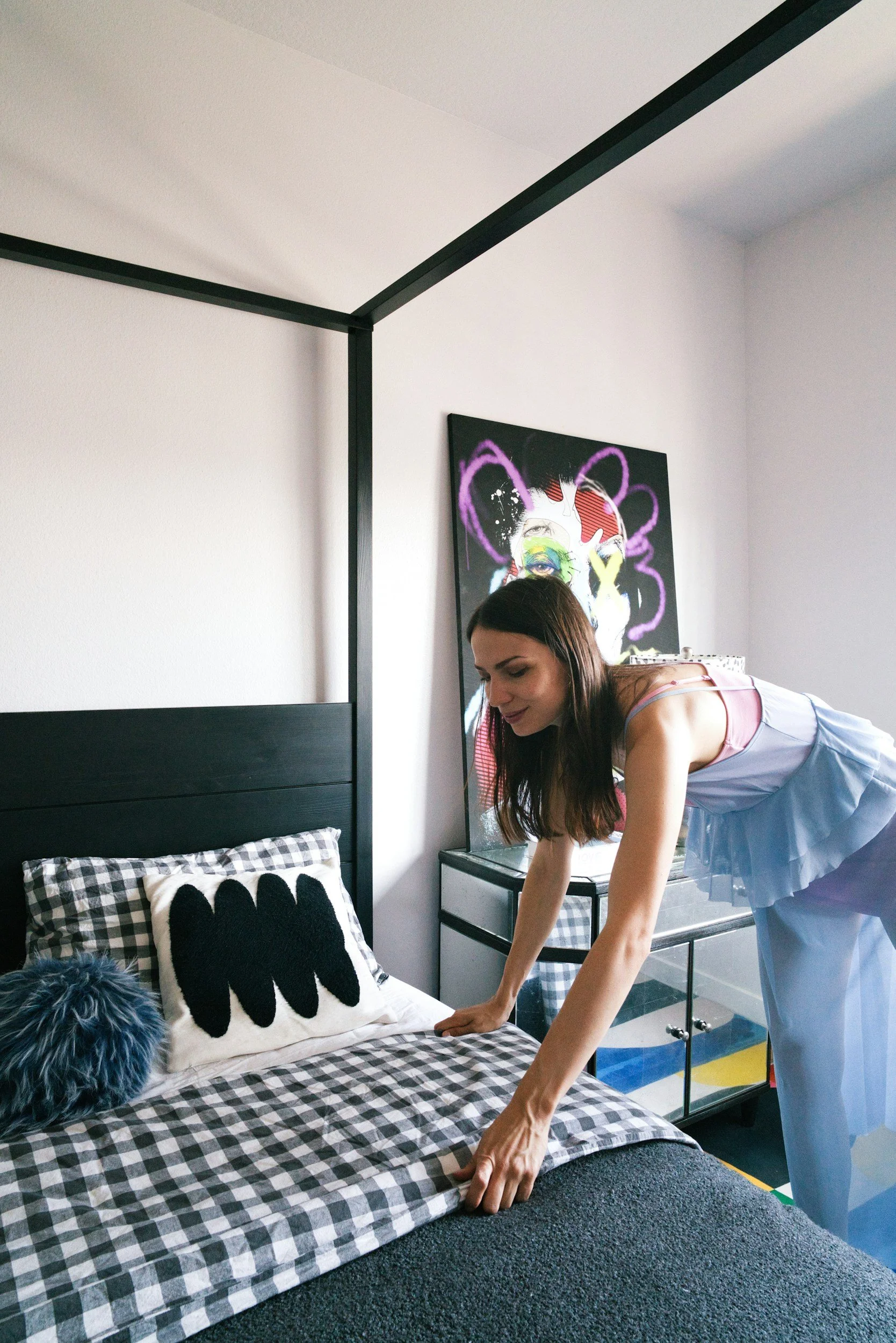 A woman with long brown hair in a pastel pink and blue nightgown making her bed in a modern room with a black headboard, checkered bedding, and a decorative pillow with black wave patterns. A colorful abstract artwork is hanging on the white wall behind her.