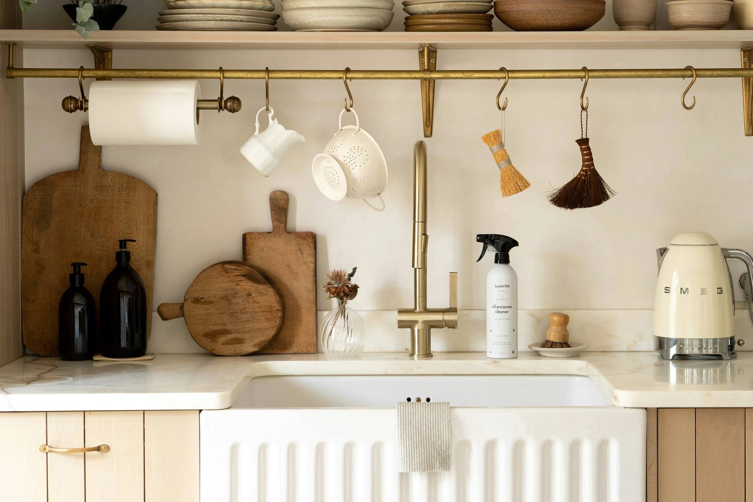 Kitchen sink with a brass faucet, surrounded by wooden cutting boards, a glass vase with dried flowers, a spray cleaner, a brush, and a small plate with a sponge. Above, a brass rod holds paper towels, cups, and cleaning brushes. Shelves above hold bowls and dishes.