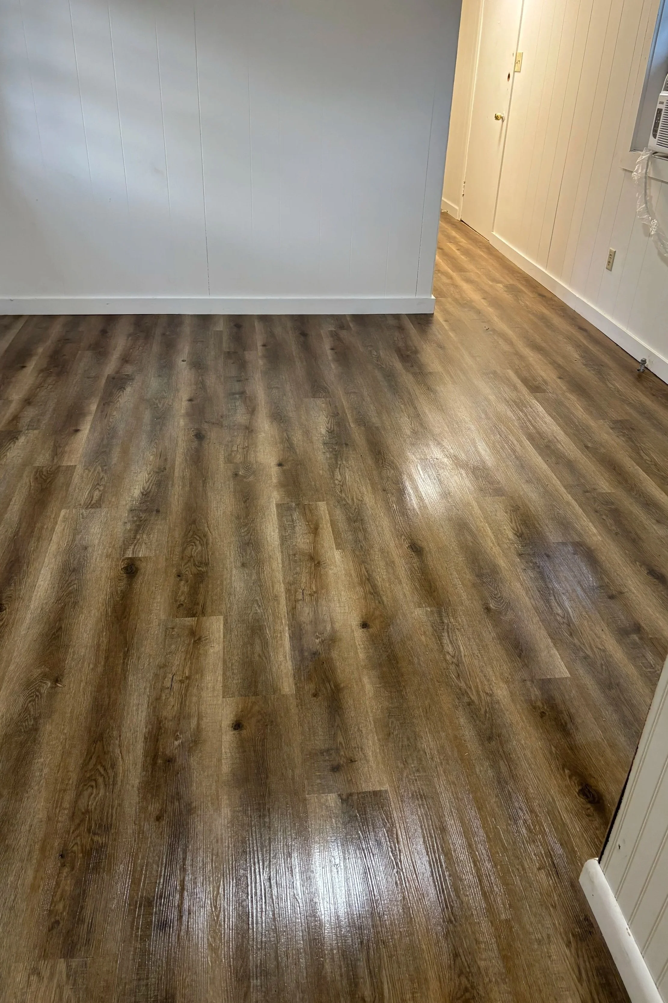 Photo of a room with newly installed brown wood laminate flooring, white paneled walls, and a doorway at the back.