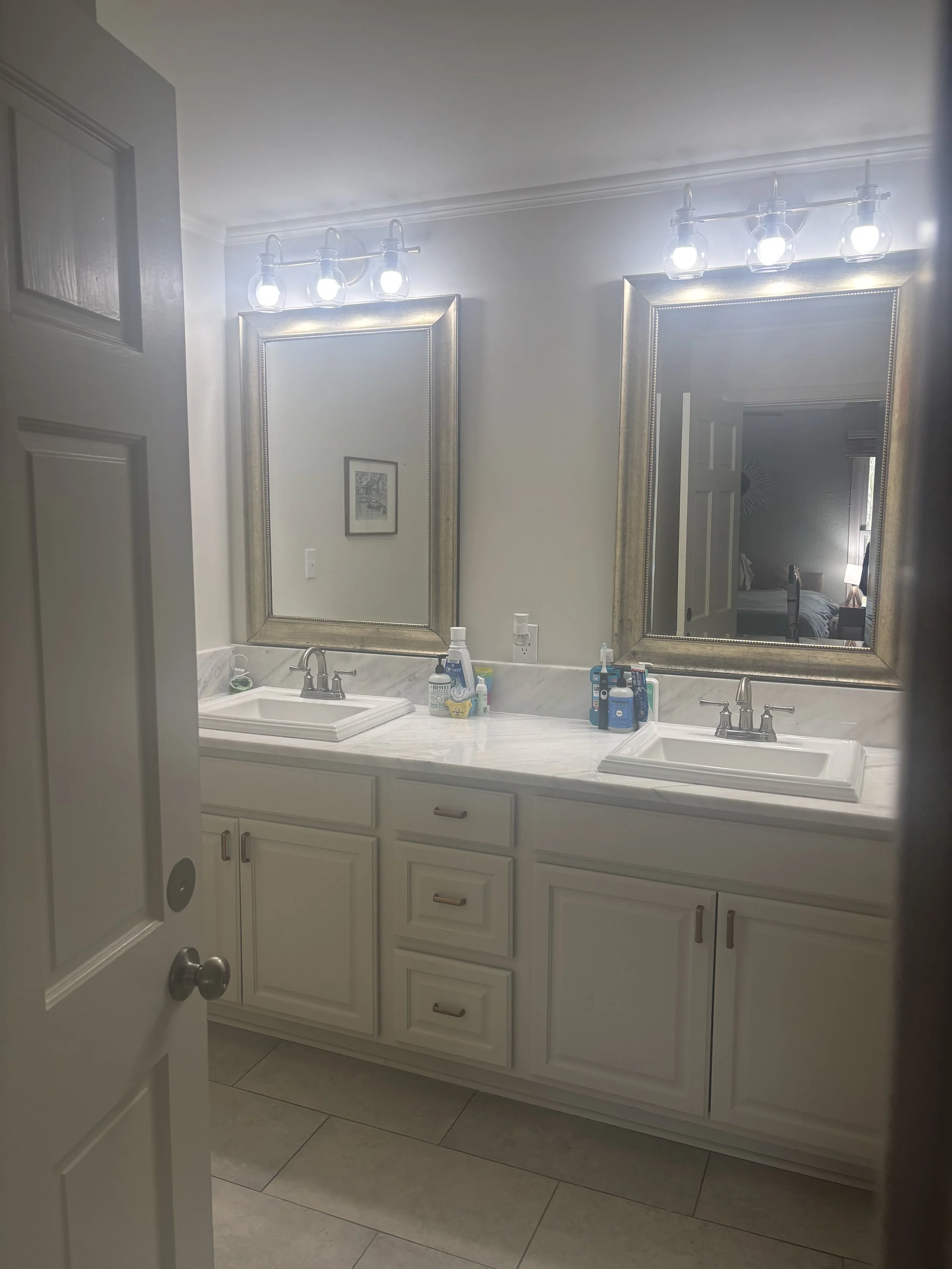 Double vanity bathroom with two mirrors, two sinks, and modern light fixtures, with toiletries on the marble countertop.