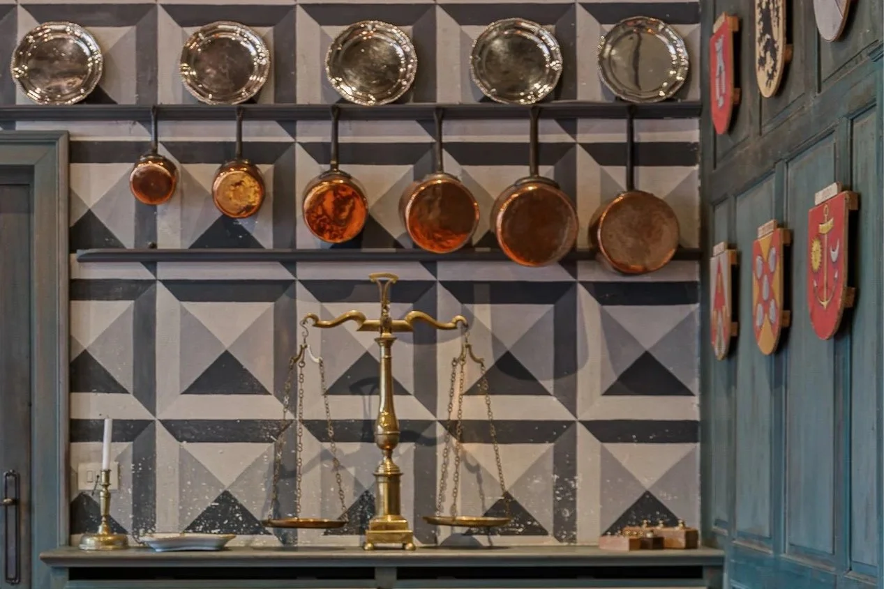 A kitchen wall decorated with hanging copper pots and stainless steel bowls above a vintage brass balance scale on a gray countertop, with a tiled black and white geometric pattern wall and wooden cabinets with colorful shields on the side.