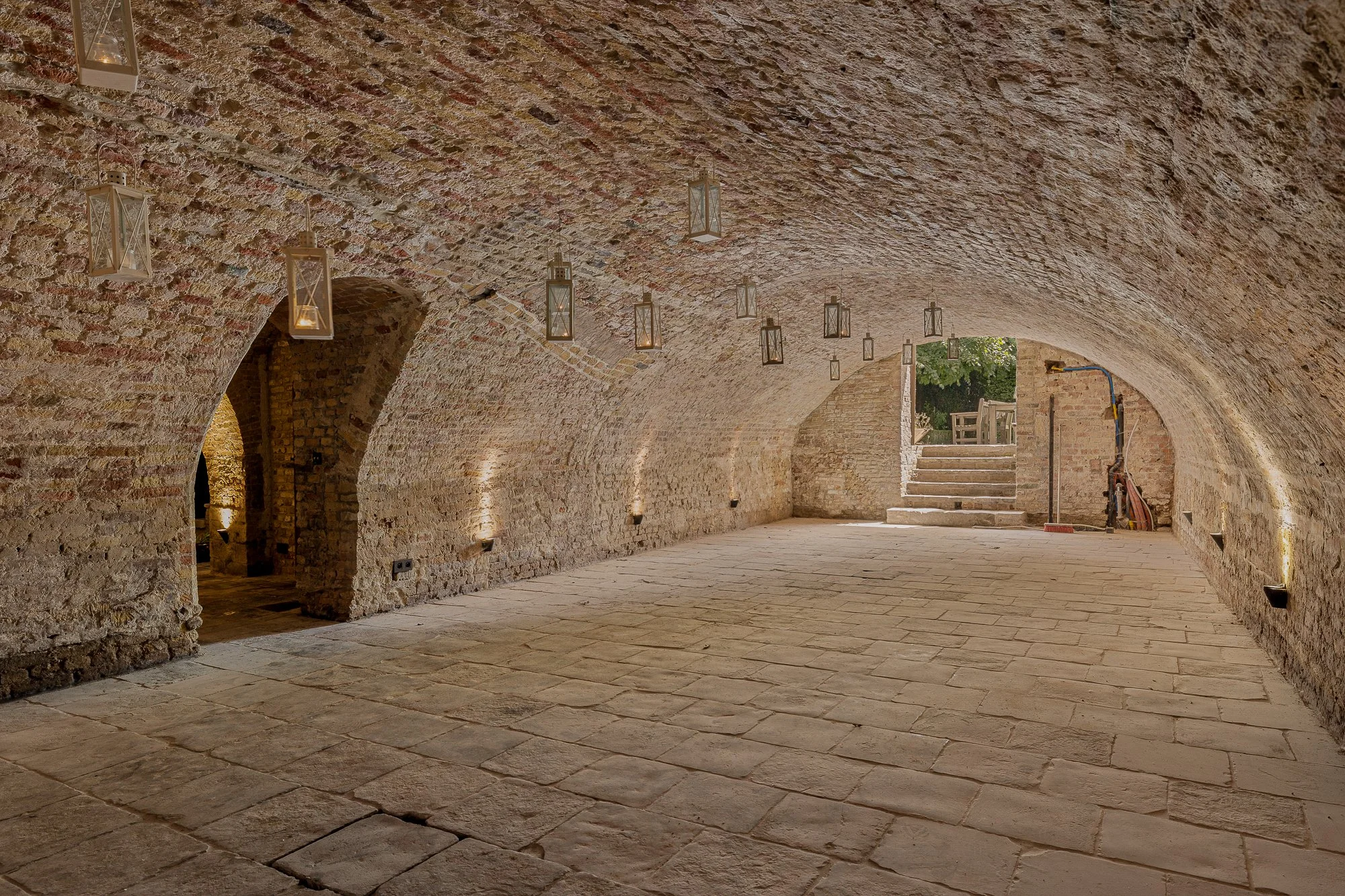 A kitchen wall decorated with hanging copper pots and stainless steel bowls above a vintage brass balance scale on a gray countertop, with a tiled black and white geometric pattern wall and wooden cabinets with colorful shields on the side.